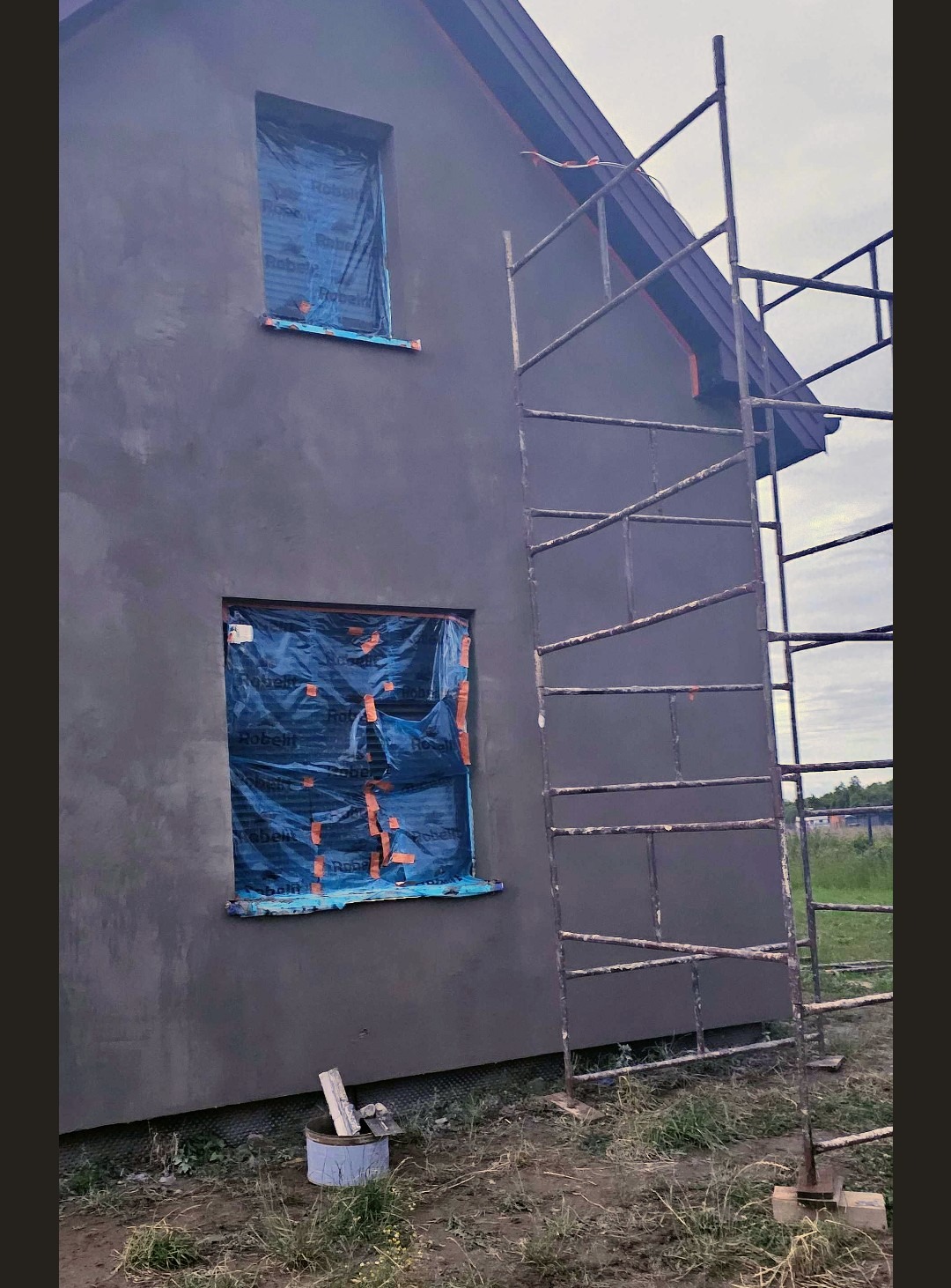 Szara elewacja domu w trakcie prac wykończeniowych. Okna zabezpieczone folią, obok rusztowanie. Widoczne narzędzia i wiadro. Świeżo nałożony tynk.