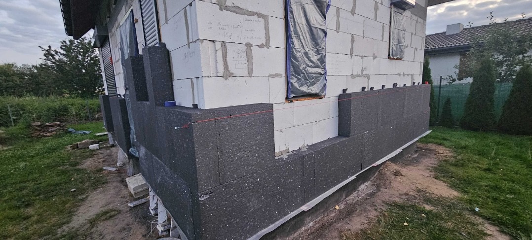 Ocieplanie ścian zewnętrznych budynku z betonu komórkowego, widoczne płyty styropianowe grafitowe, sznurek traserski i okna zabezpieczone folią.