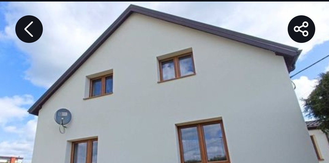 Dwukondygnacyjny dom z jasną elewacją i brązowymi ramami okiennymi. Na ścianie widoczna antena satelitarna. Ujęcie z dołu, niebo z chmurami w tle.