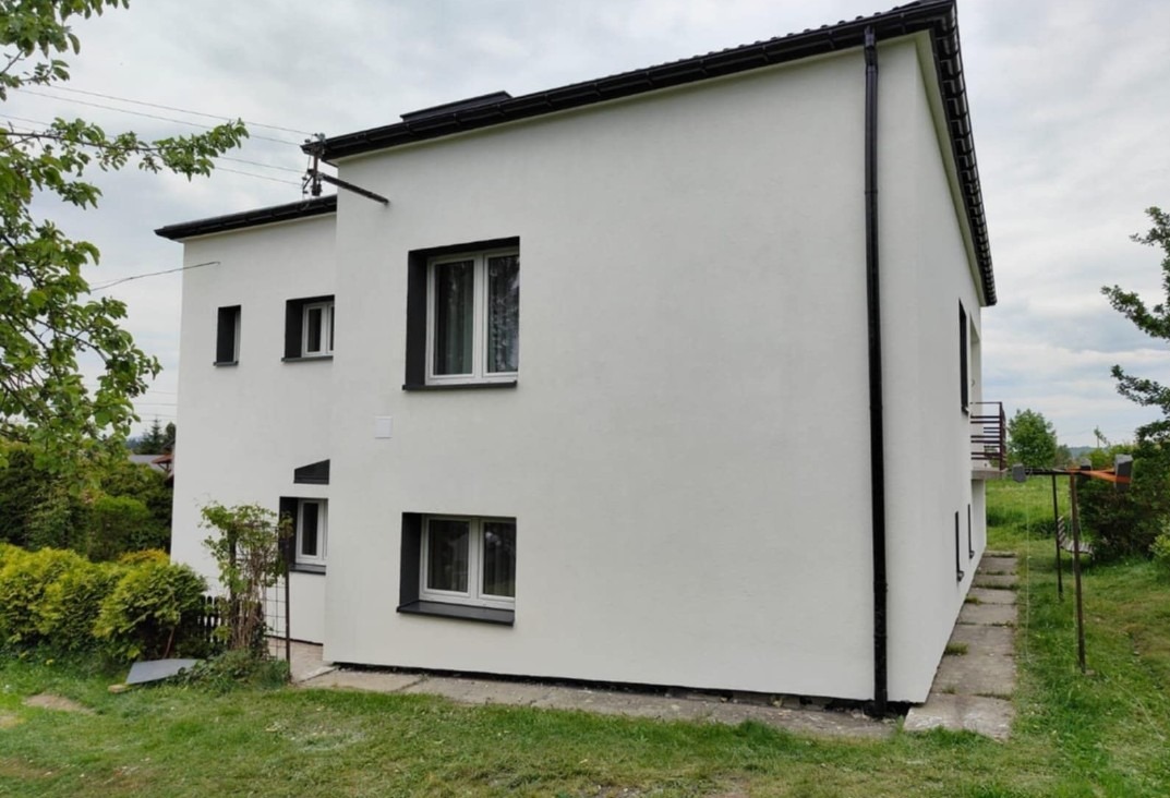 Biały, dwukondygnacyjny dom z czarnymi ramami okiennymi i dachem, otoczony zielenią. Widok z boku, ukazujący elewację i fragment ogrodu. Minimalistyczny styl.