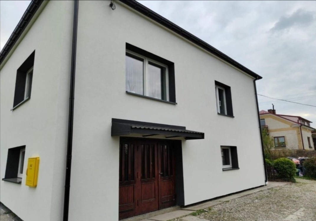 Elewacja domu po remoncie: biała ściana, ciemne obramowania okien i drzwi, żółta skrzynka pocztowa. Widok z dołu, pochmurne niebo w tle.