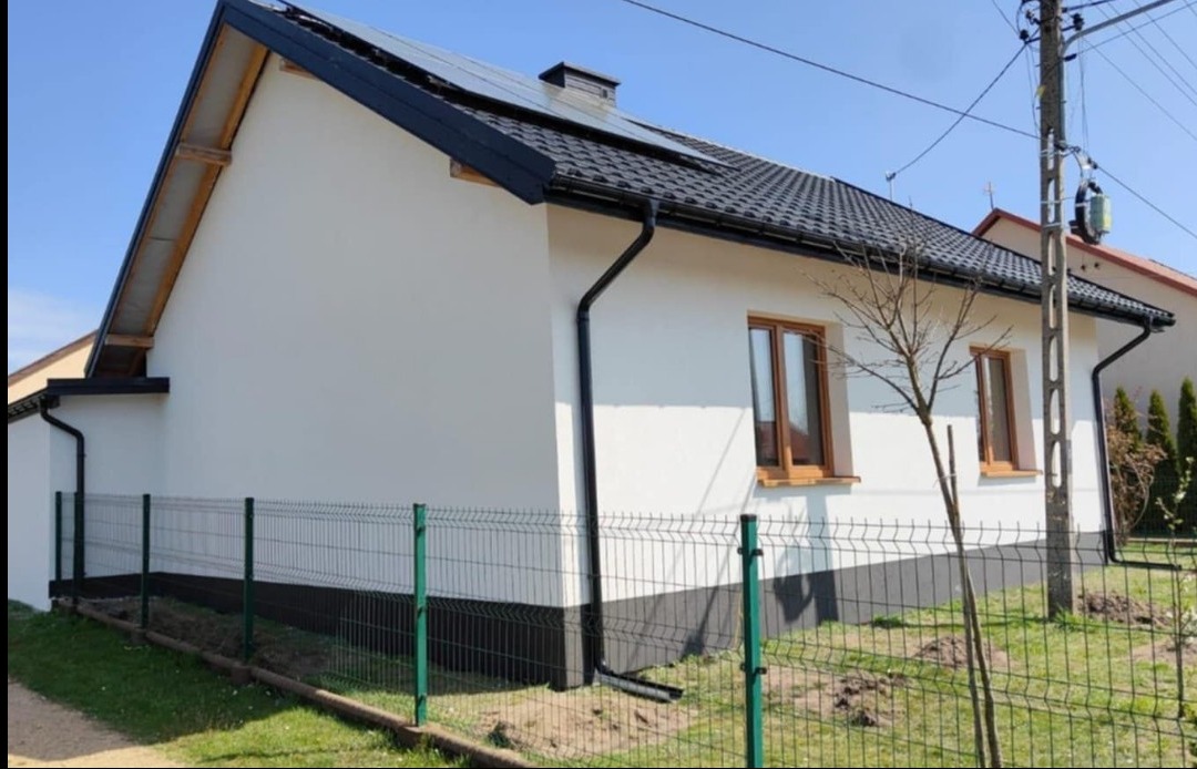 Boczny widok domu po remoncie elewacji, z czarnym dachem i rynnami, ogrodzony siatką. Widoczne okna z brązowymi ramami i fragment ogrodu.