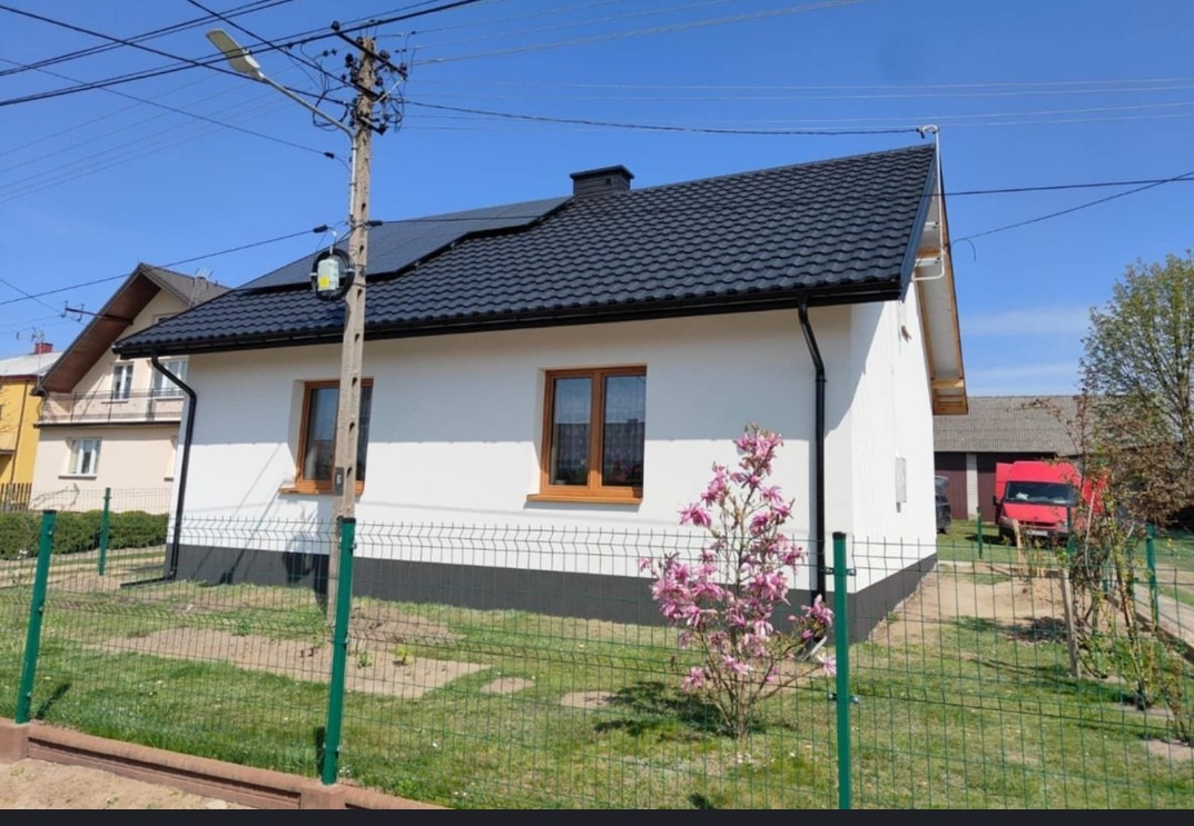 Biały dom z ciemnym dachem i brązowymi oknami, otoczony zielonym płotem i kwitnącą magnolią. Widoczny słup energetyczny i czerwony samochód w tle.