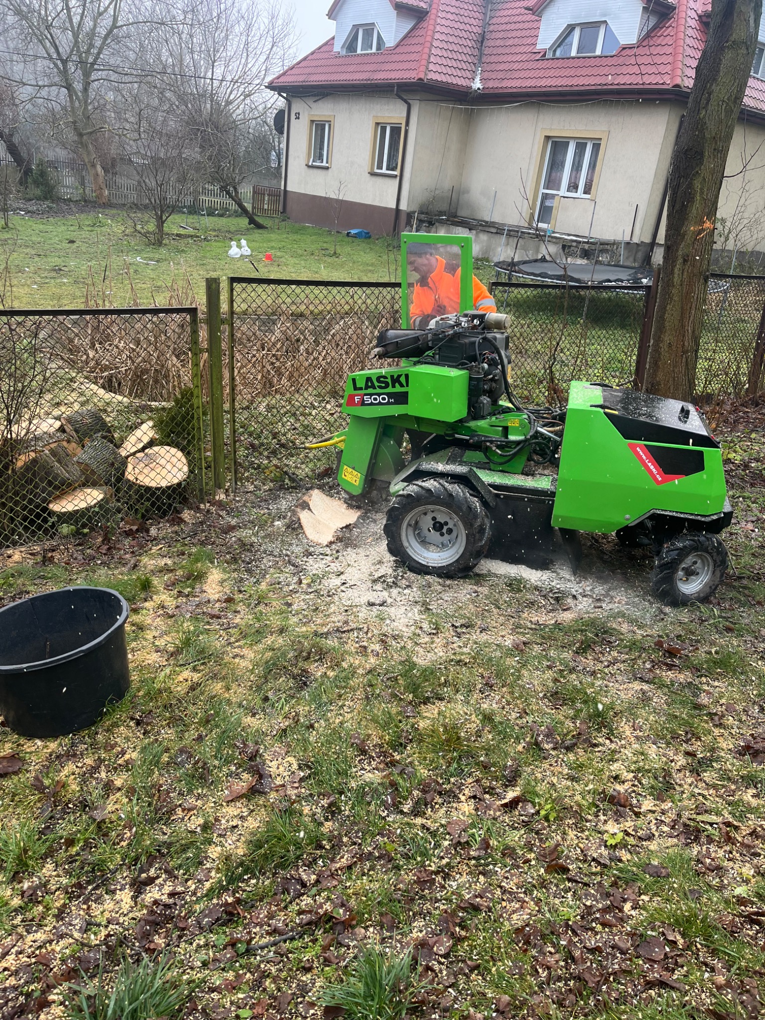Zielona frezarka do pni drzew LASKO F500H w trakcie pracy, w tle stos pociętych pni i dom z czerwonym dachem. Wokół maszyny widać wióry.