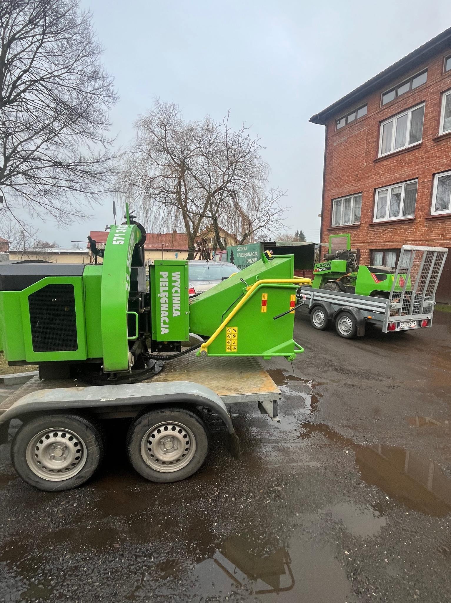 Dwie przyczepy z maszynami do pielęgnacji drzew, w tym rębakiem do gałęzi z napisem 'Wycinka Pielęgnacja', stoją na mokrym placu przed budynkiem.