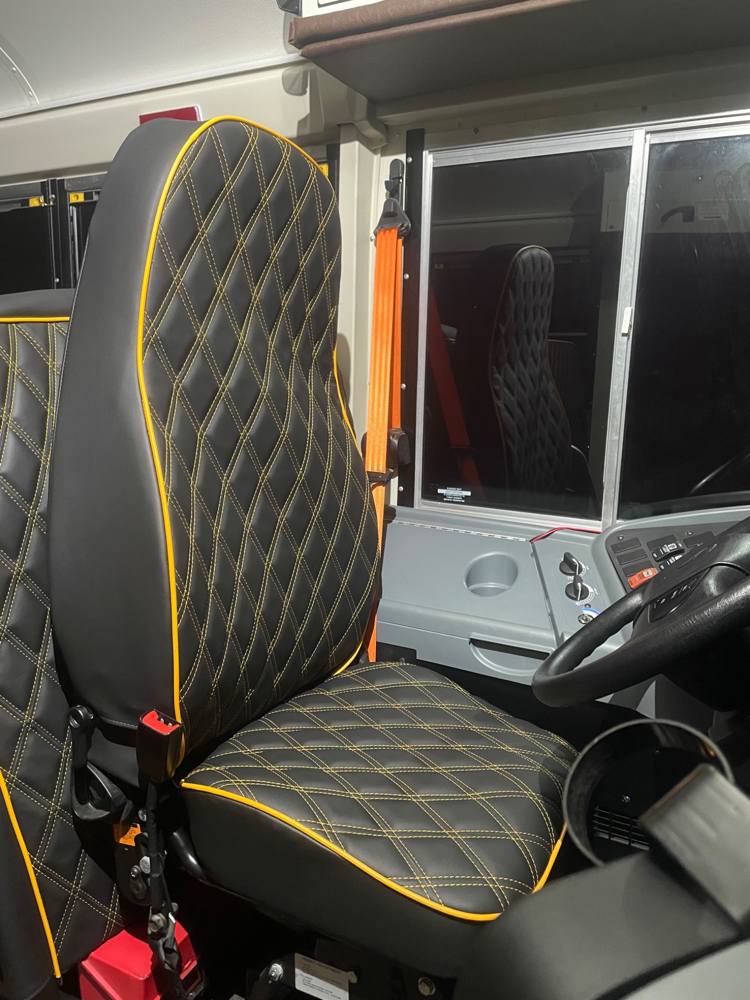 Czarne, pikowane siedzisko z żółtą lamówką w samochodzie dostawczym. Widoczny fragment deski rozdzielczej i pomarańczowy pas bezpieczeństwa.