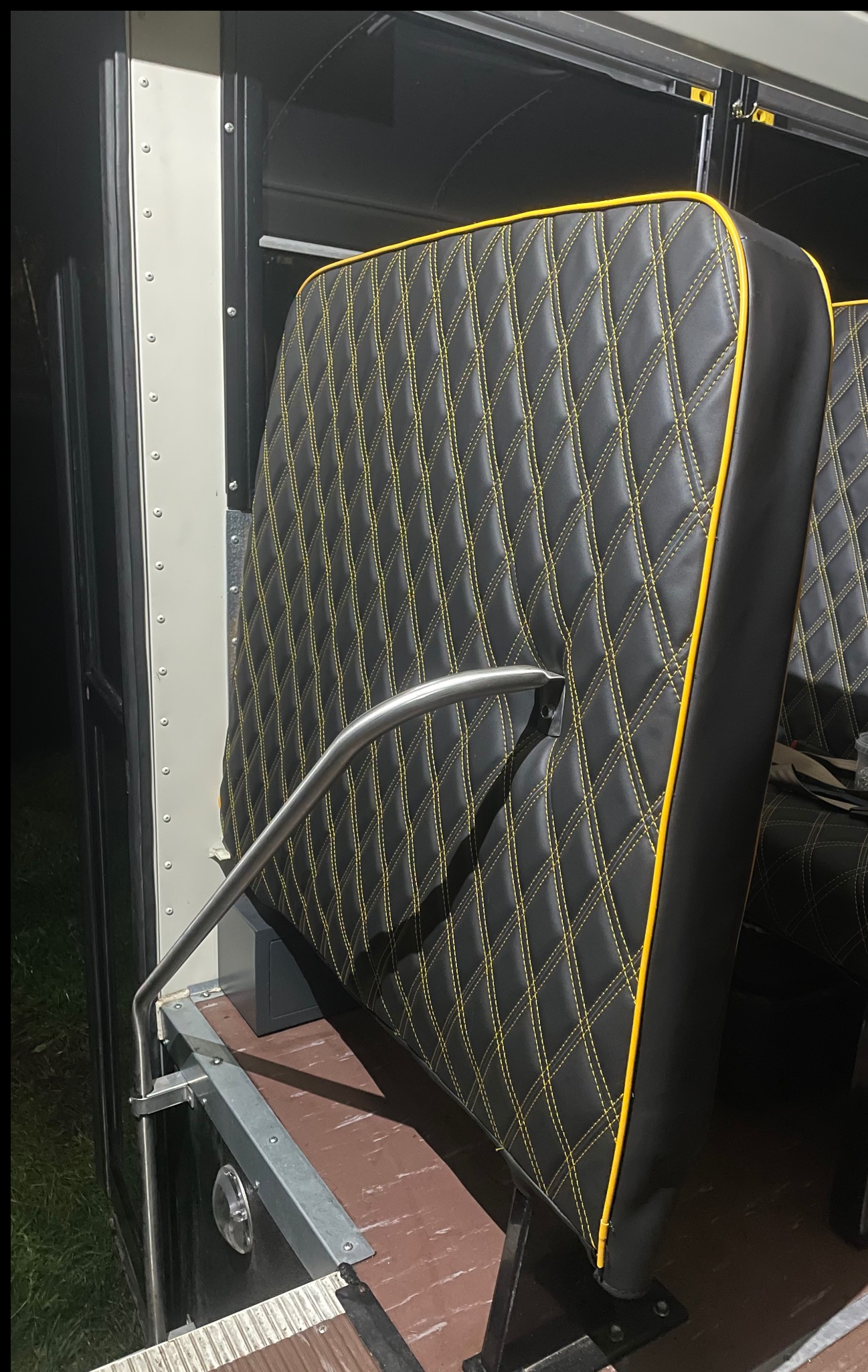 Czarne, pikowane siedzisko z żółtą lamówką w autobusie, widoczne schodki i poręcz. Detal tapicerki z wyraźnym wzorem rombów i kontrastowym obszyciem.