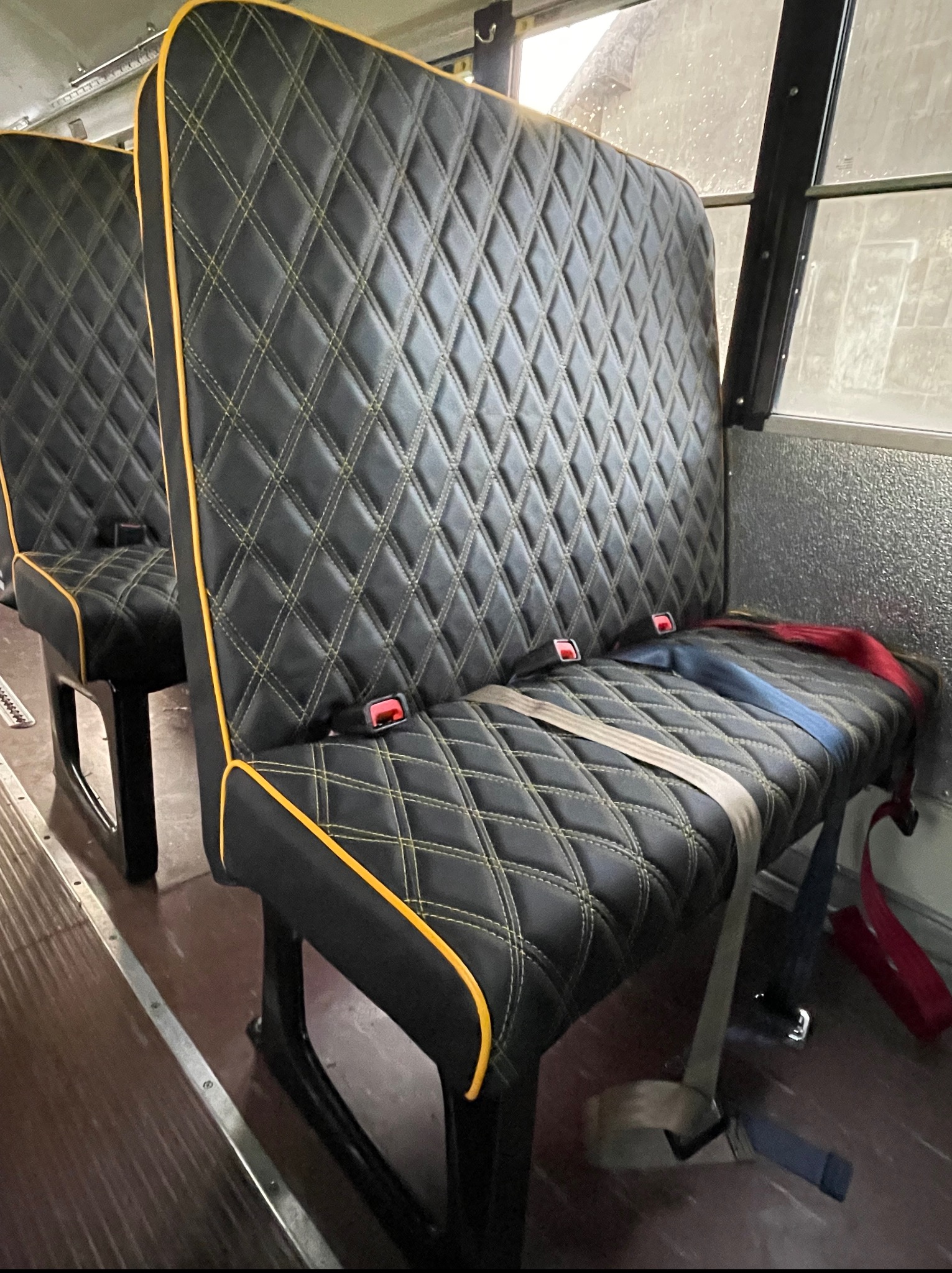Czarne, pikowane siedzenie z żółtą lamówką w pojeździe. Widoczne pasy bezpieczeństwa i metalowe wykończenie wnętrza. Detal tapicerki samochodowej.