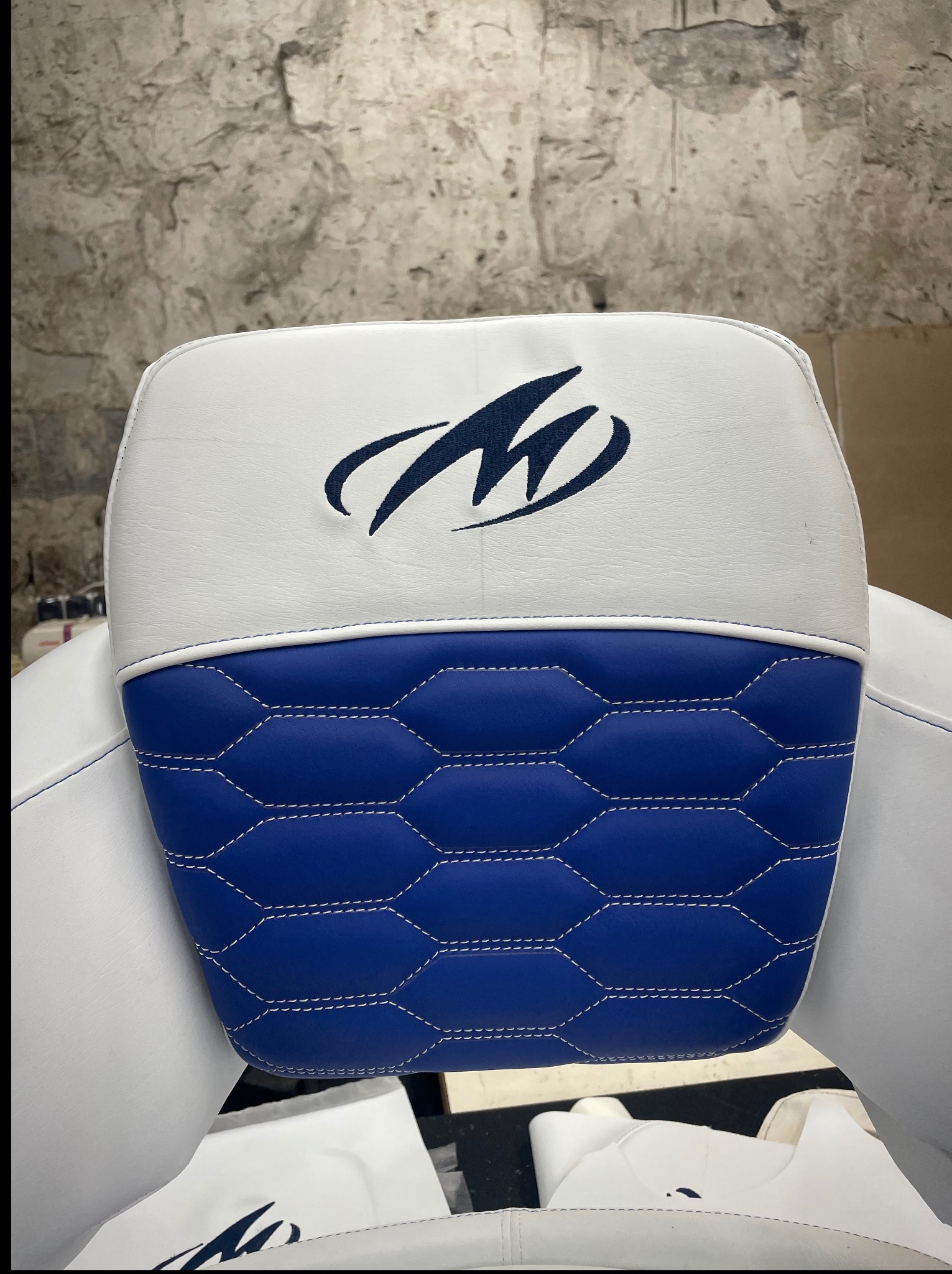 Biało-niebieski zagłówek samochodowy z logo, wykończony pikowaniem i białymi szwami, na tle surowej ściany. Detal tapicerki po renowacji.