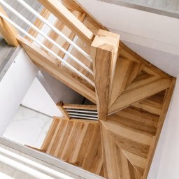 KCR - Spiralne schody z jasnego drewna, widok z góry. Białe balustrady i szare panele podłogowe. Nietypowy układ stopni tworzy geometryczny wzór.