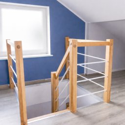KCR - Jasne drewniane schody z białymi, metalowymi prętami balustrady, widziane z góry, na tle błękitnej ściany i poddasza. Minimalistyczny design.