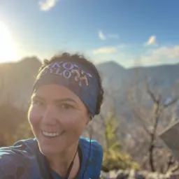 Uśmiechnięta kobieta w sportowej opasce na tle gór i błękitnego nieba. Widoczny znak ostrzegawczy. Słońce oświetla twarz, tworząc ciepły blask.