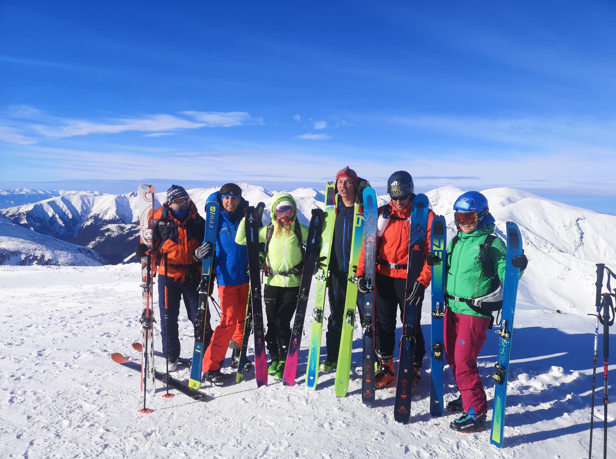 Grupa narciarzy skiturowych na tle ośnieżonych gór w słoneczny dzień. Różnokolorowe kurtki i narty. Widok na szczyty górskie pokryte śniegiem.