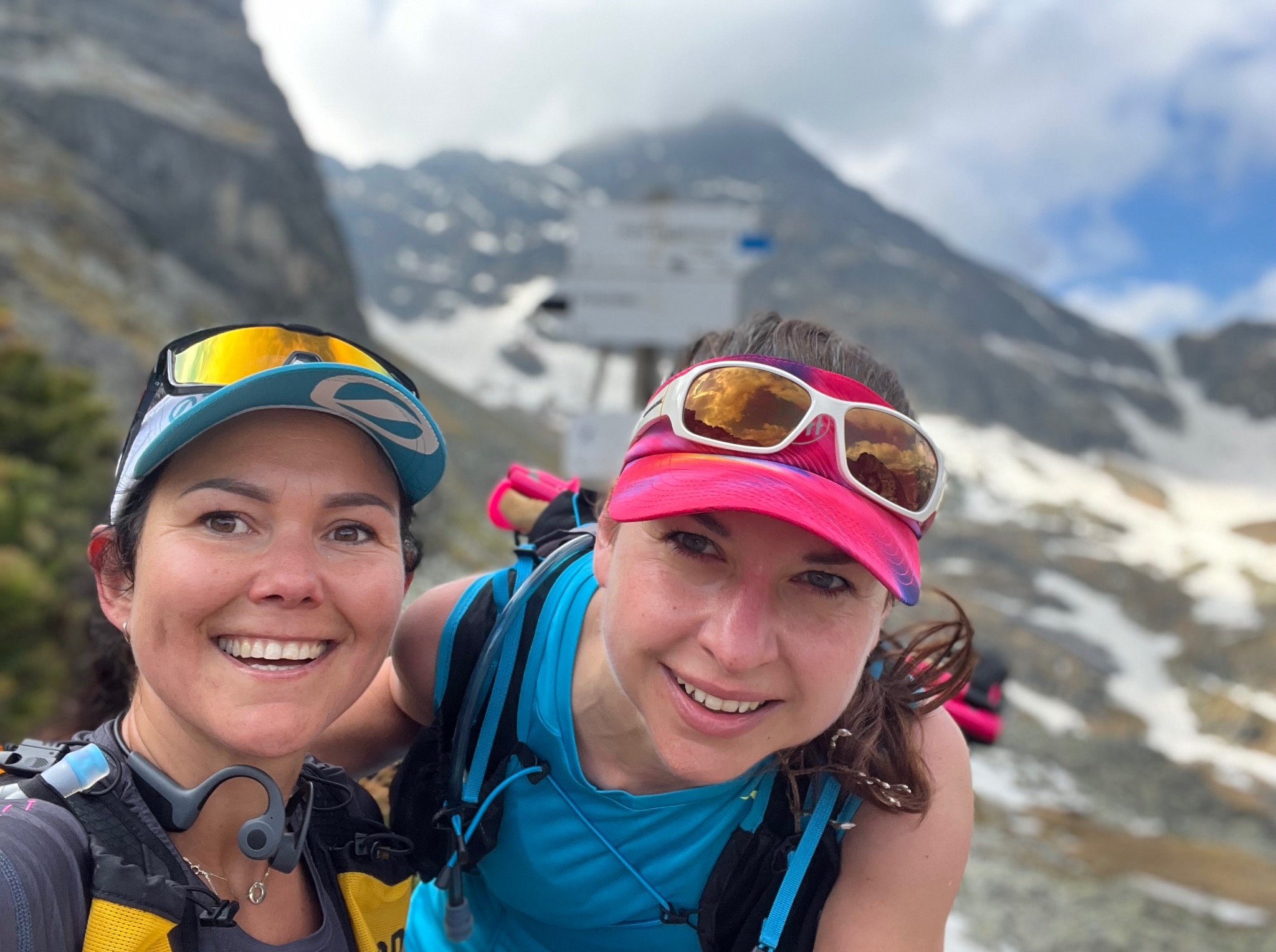 Uśmiechnięte kobiety w sportowych strojach, robiące selfie na tle górskiego krajobrazu z tabliczkami szlaków. Widoczne góry, śnieg i błękitne niebo.