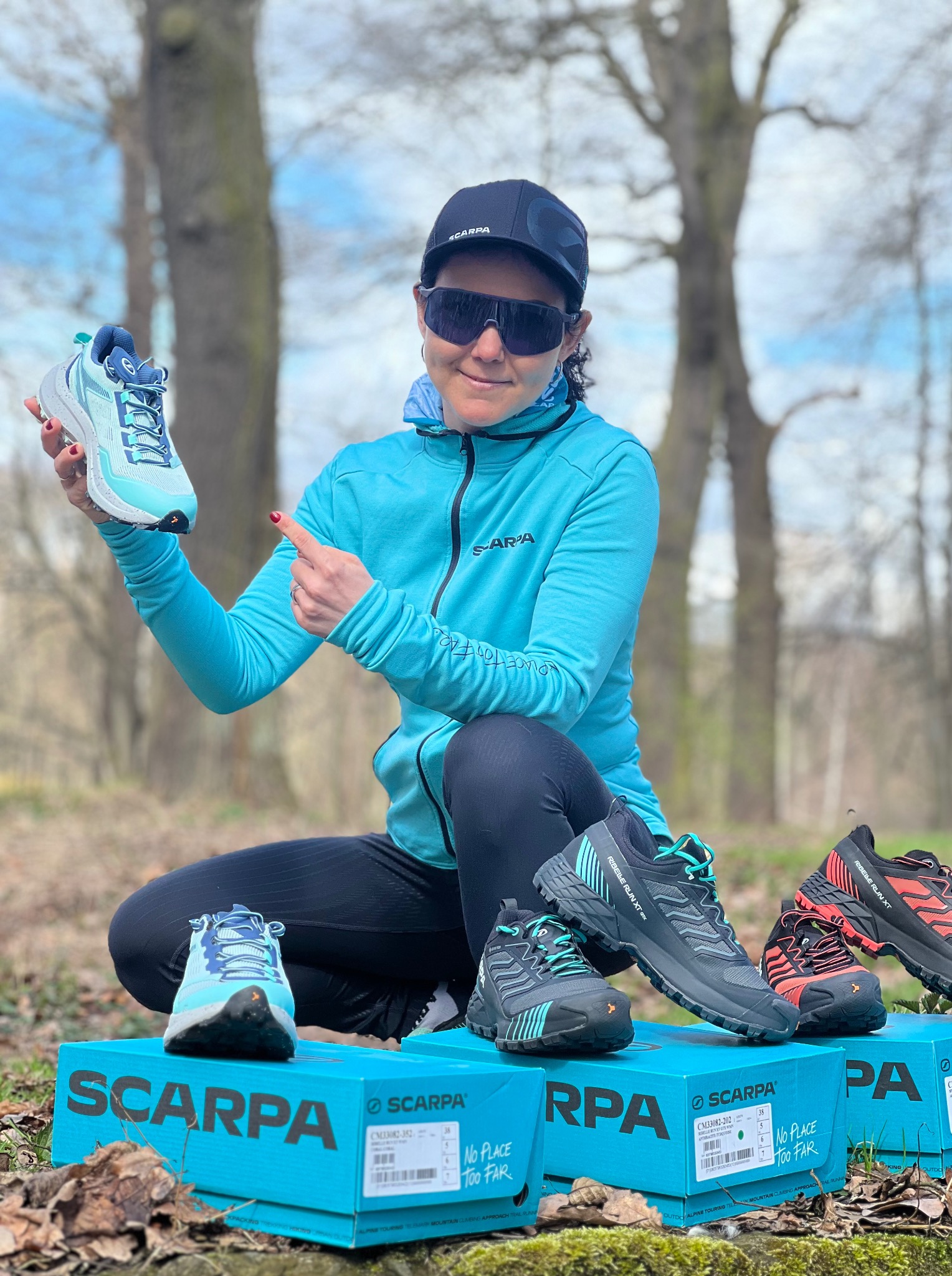 Kobieta w sportowym stroju prezentuje buty do biegania Scarpa na tle leśnym, siedząc na pudełkach z obuwiem. Uśmiechnięta, w okularach przeciwsłonecznych.