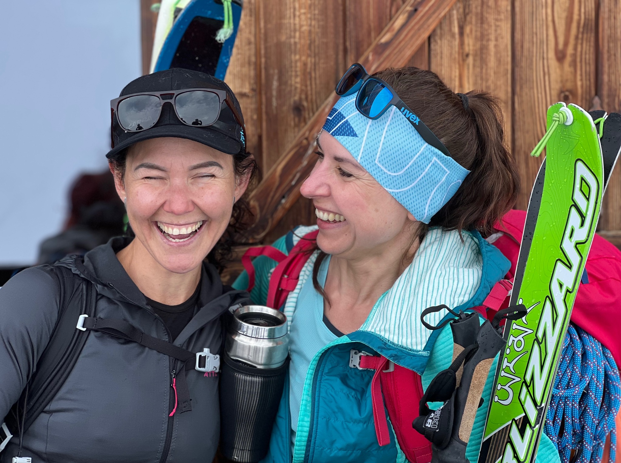 Dwie uśmiechnięte kobiety w sportowych strojach, z nartami i kijkami, prawdopodobnie po treningu biegowym w górach. Widoczne detale sprzętu i górskie tło.