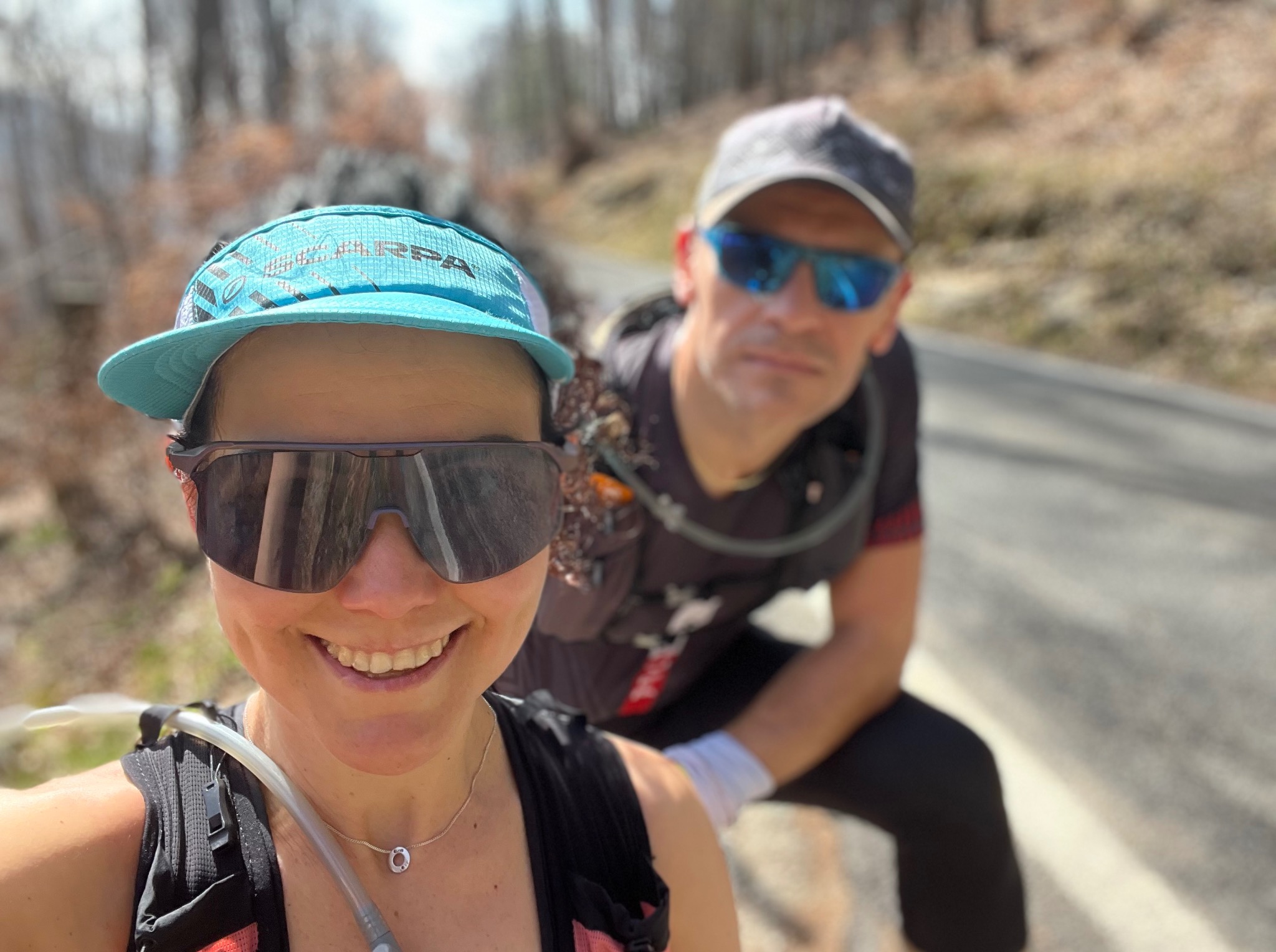 Uśmiechnięta kobieta w okularach i czapce biegowej, robiąca selfie z mężczyzną w tle, podczas treningu biegowego w górach. Widoczna trasa i leśne otoczenie.
