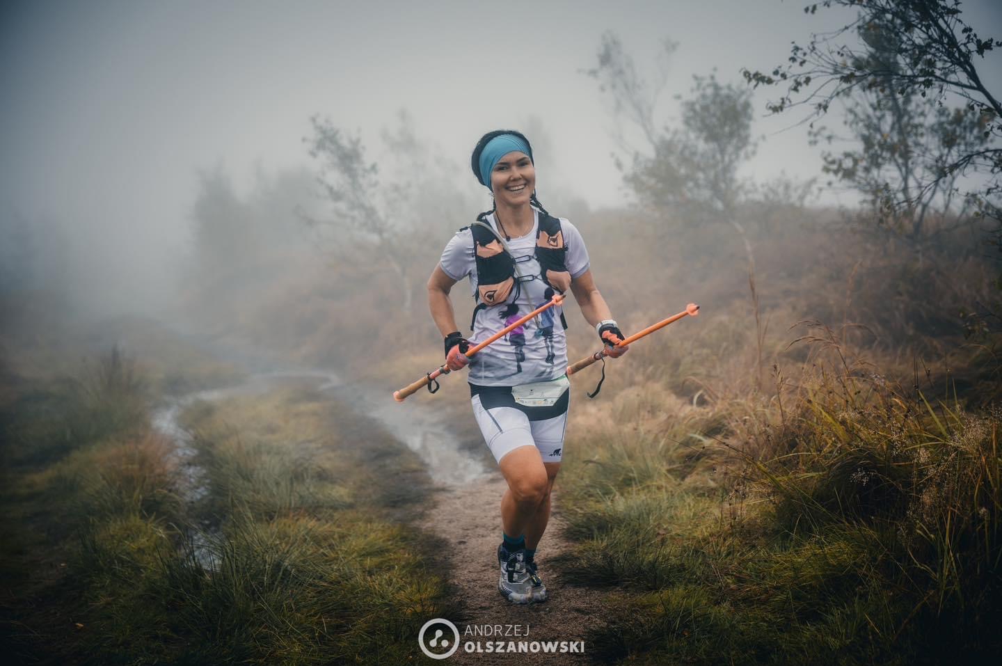 Uśmiechnięta kobieta biegnie z kijkami w mglistym terenie. Widoczny strój sportowy, plecak biegowy i opaska na głowie. Dynamiczne ujęcie w naturalnym otoczeniu.