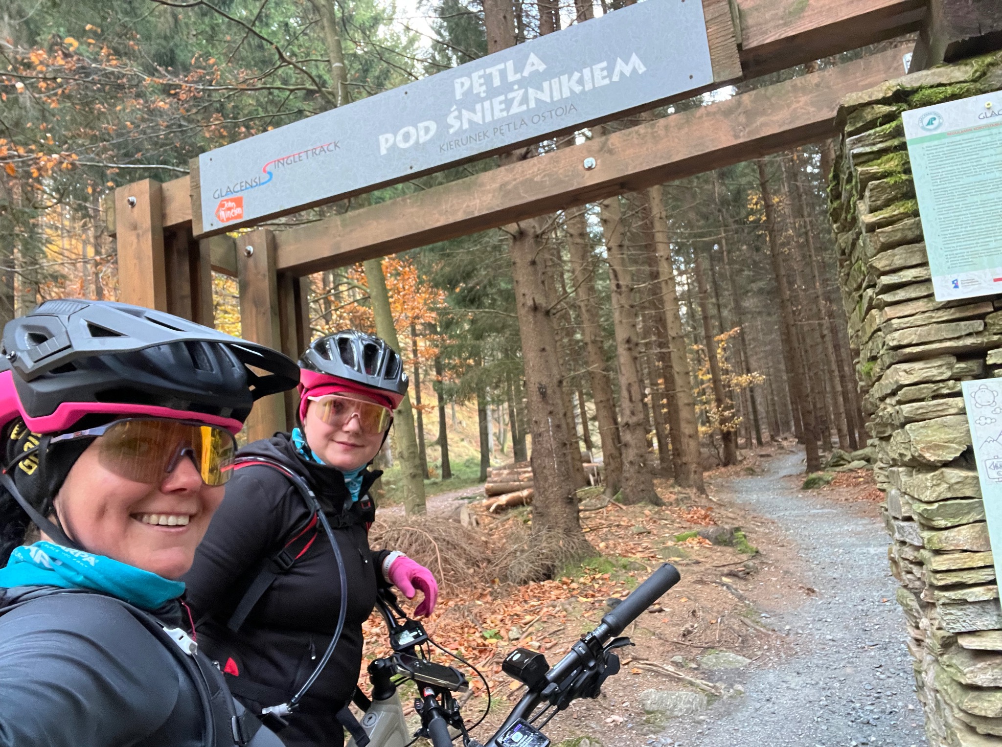Dwie uśmiechnięte kobiety w kaskach rowerowych i okularach na tle leśnej trasy rowerowej 'Pętla pod Śnieżnikiem'. Jesienna aura, widoczne drzewa i ścieżka.