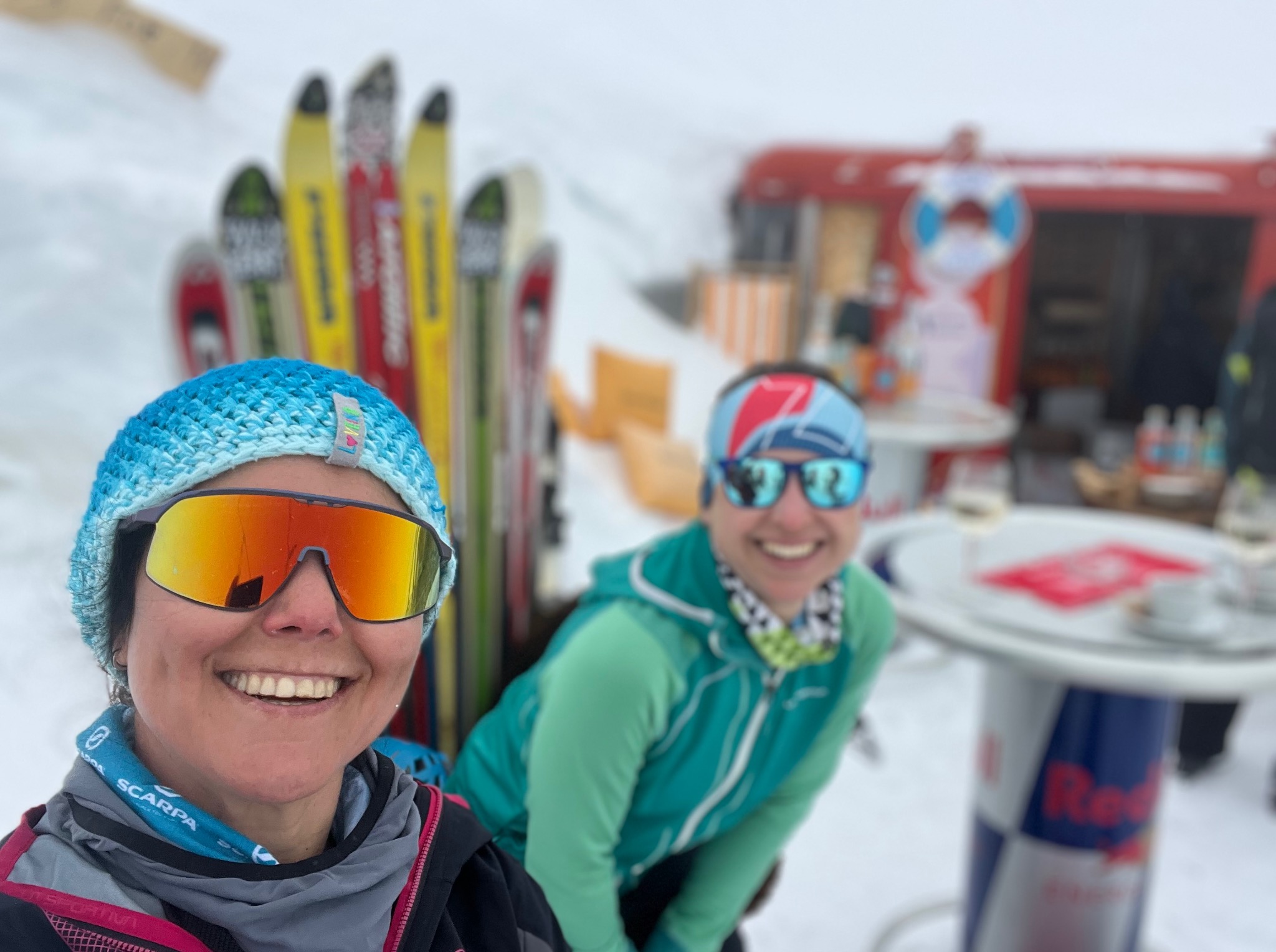 Uśmiechnięte kobiety w sportowych okularach na tle zaśnieżonego stoku z nartami i barem, prawdopodobnie po treningu biegowym.