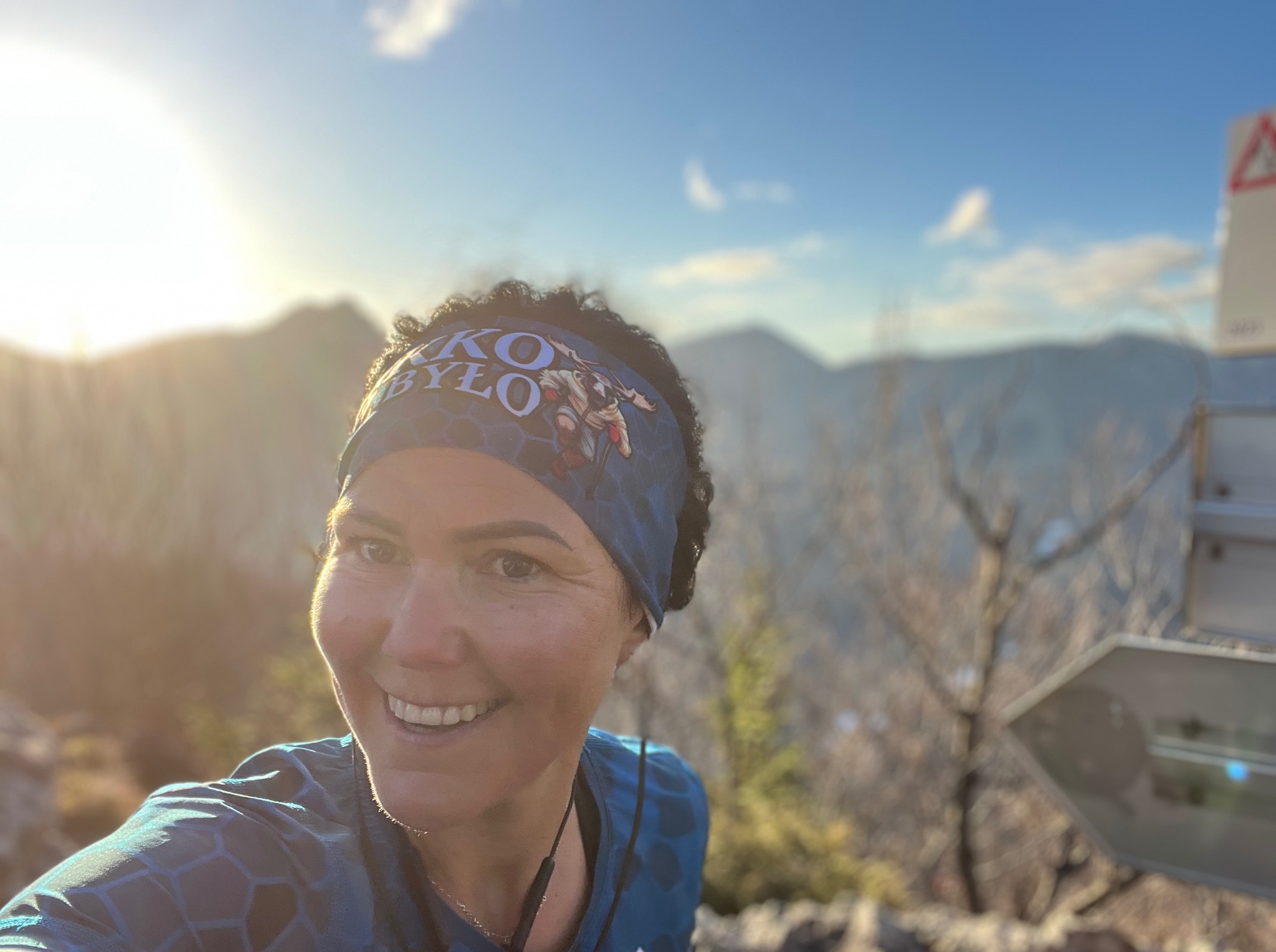 Uśmiechnięta kobieta w sportowej opasce na tle gór i błękitnego nieba. Widoczny znak ostrzegawczy. Słońce oświetla twarz, tworząc ciepły blask.