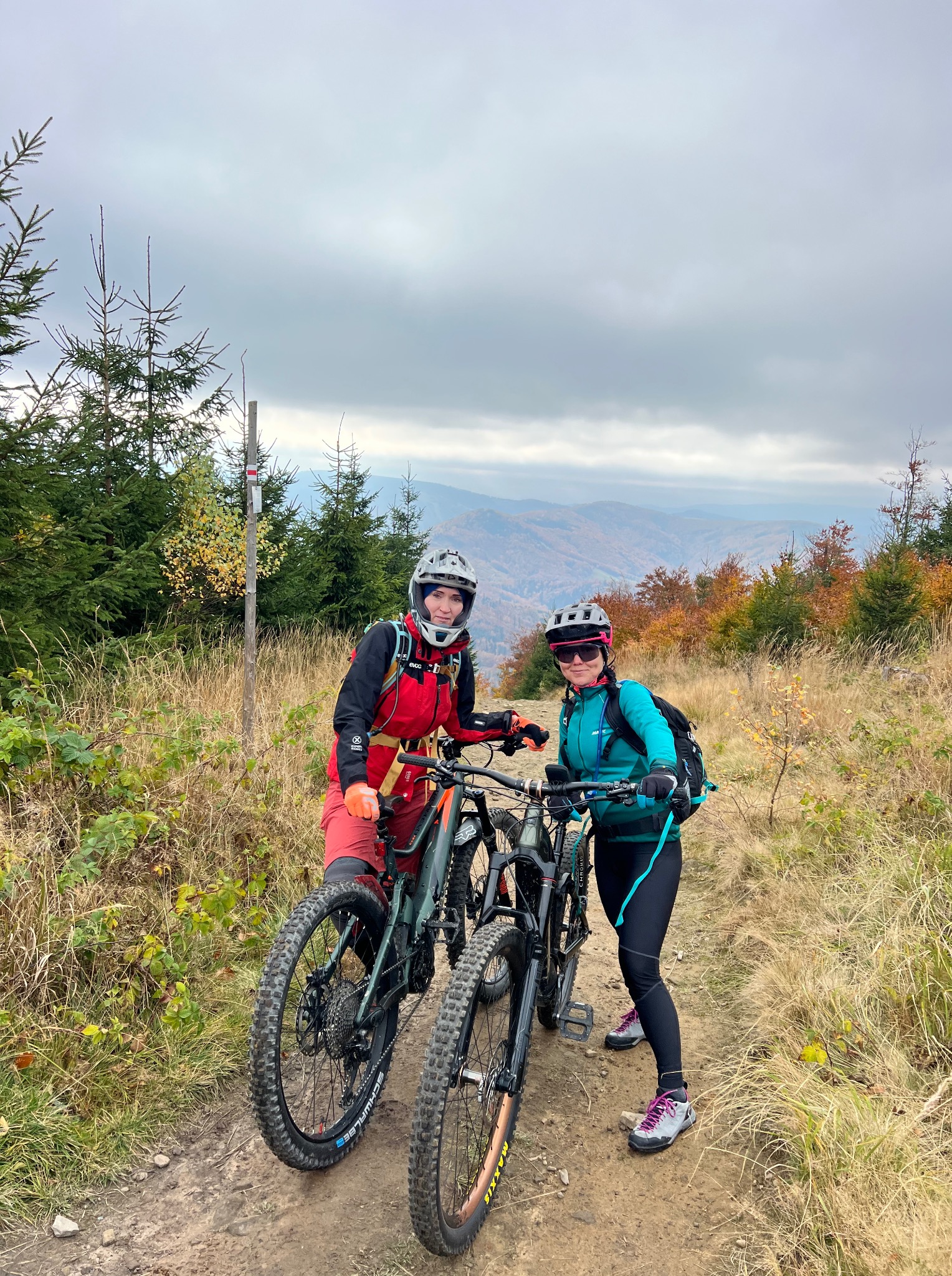 Dwie kobiety w kaskach i strojach sportowych z rowerami górskimi na szlaku. W tle góry i las w jesiennych barwach. Aktywny wypoczynek na łonie natury.