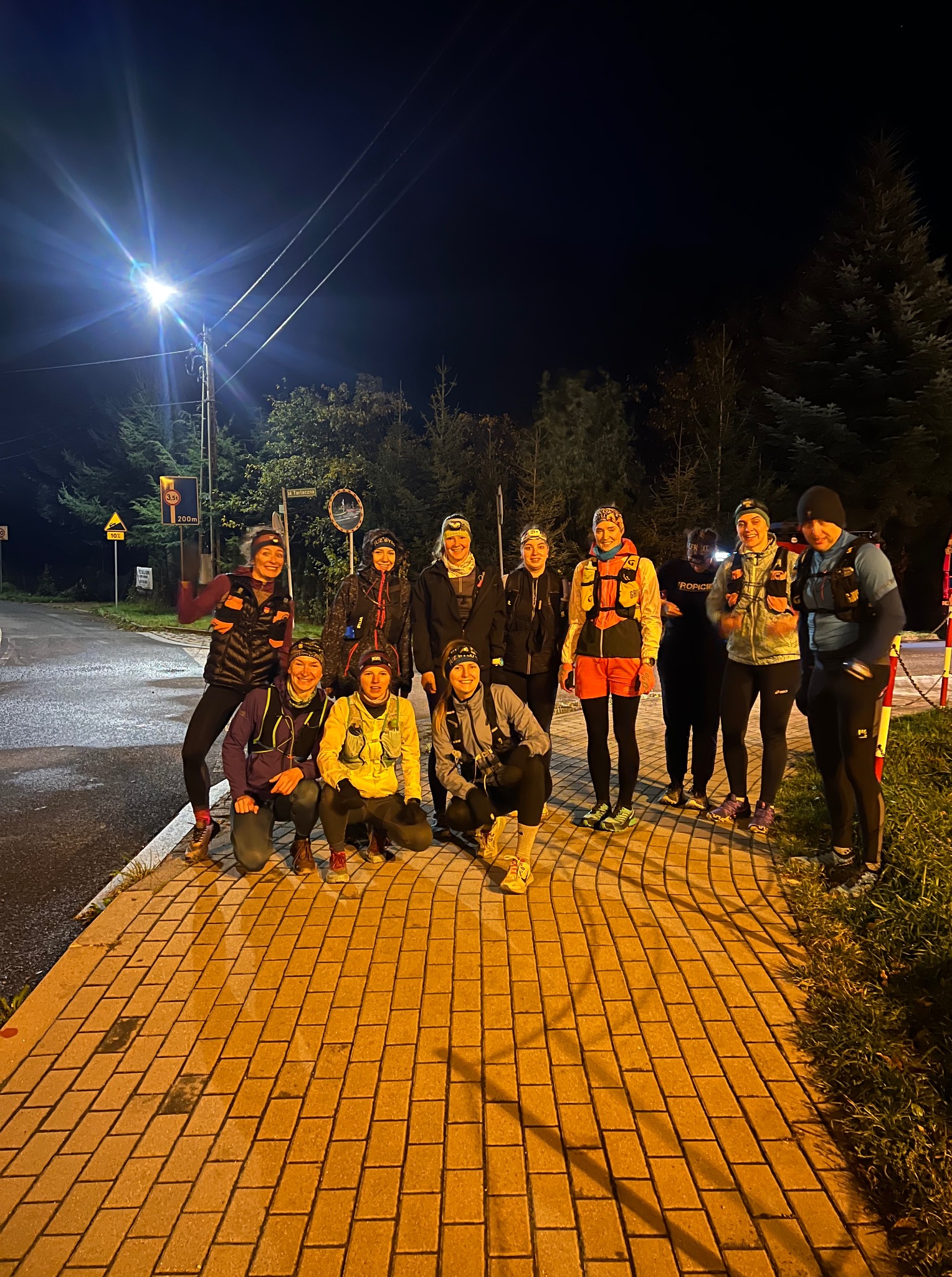 Grupa biegaczek w strojach sportowych pozuje na chodniku w nocy, oświetlona latarnią. Widoczne znaki drogowe i drzewa w tle. Uczestniczki biegu nocnego.