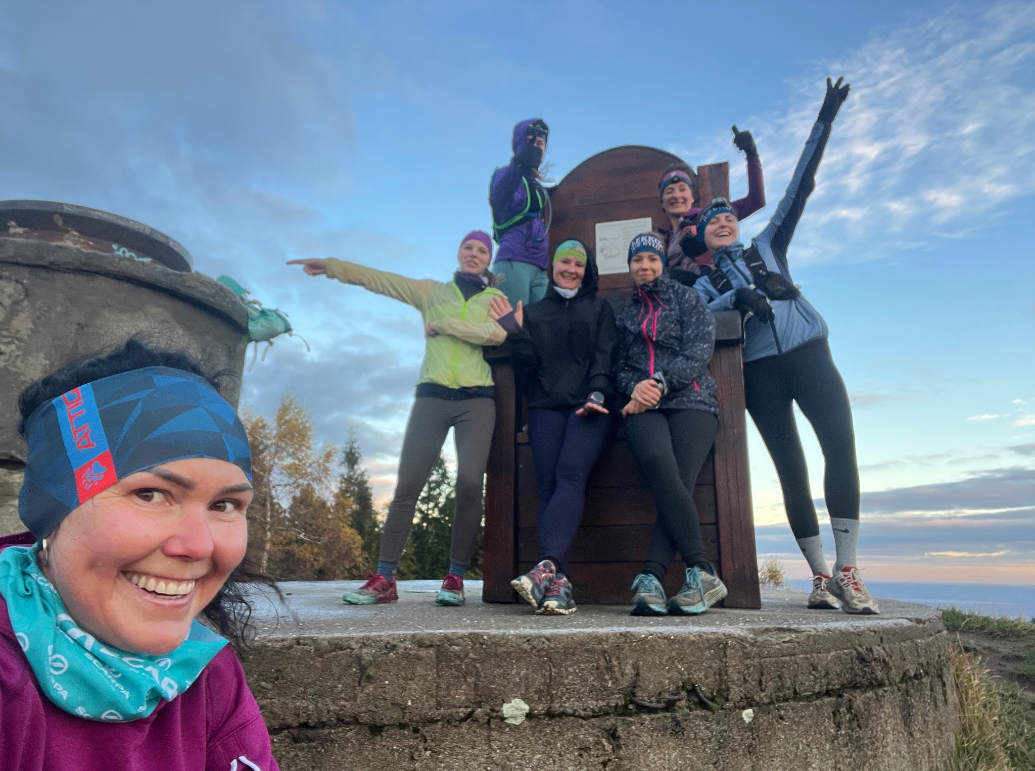 Grupa uśmiechniętych kobiet w sportowych strojach pozuje na tle krajobrazu podczas treningu biegowego. Widoczny betonowy bunkier i drewniana tablica informacyjna.