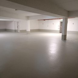 Epoxy Floor - Jasnoszara, gładka posadzka w przestronnym, pustym pomieszczeniu z betonowymi filarami i numerami na ścianach. Widoczne drzwi ewakuacyjne.