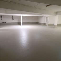 Epoxy Floor - Jasnoszara, gładka posadzka w garażu podziemnym z numerowanymi miejscami parkingowymi. Widoczne słupy i fragment drzwi.