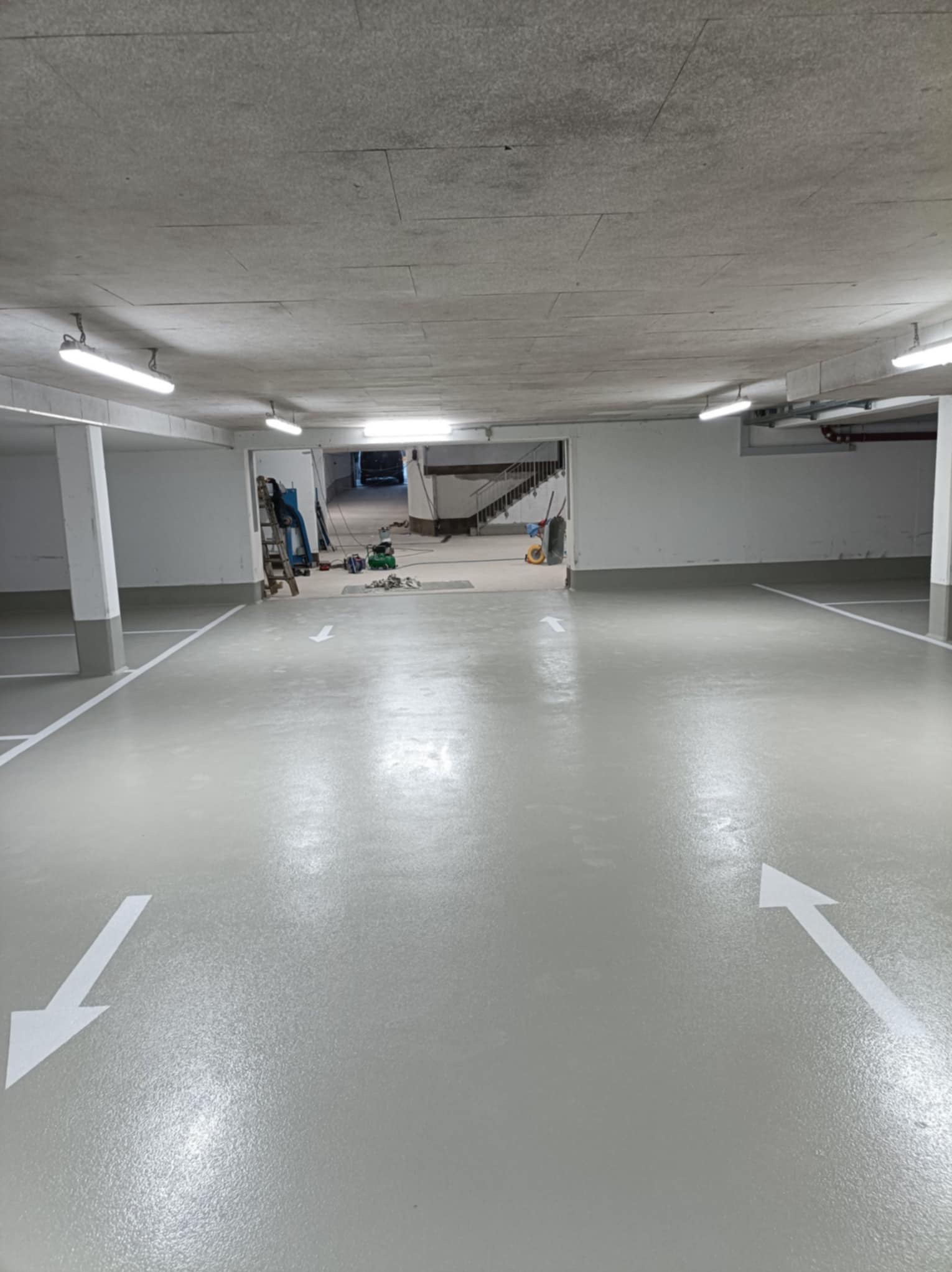 Jasnoszara posadzka w garażu podziemnym z białymi strzałkami kierunkowymi. W tle widoczne narzędzia i drabina, sugerujące prace wykończeniowe.