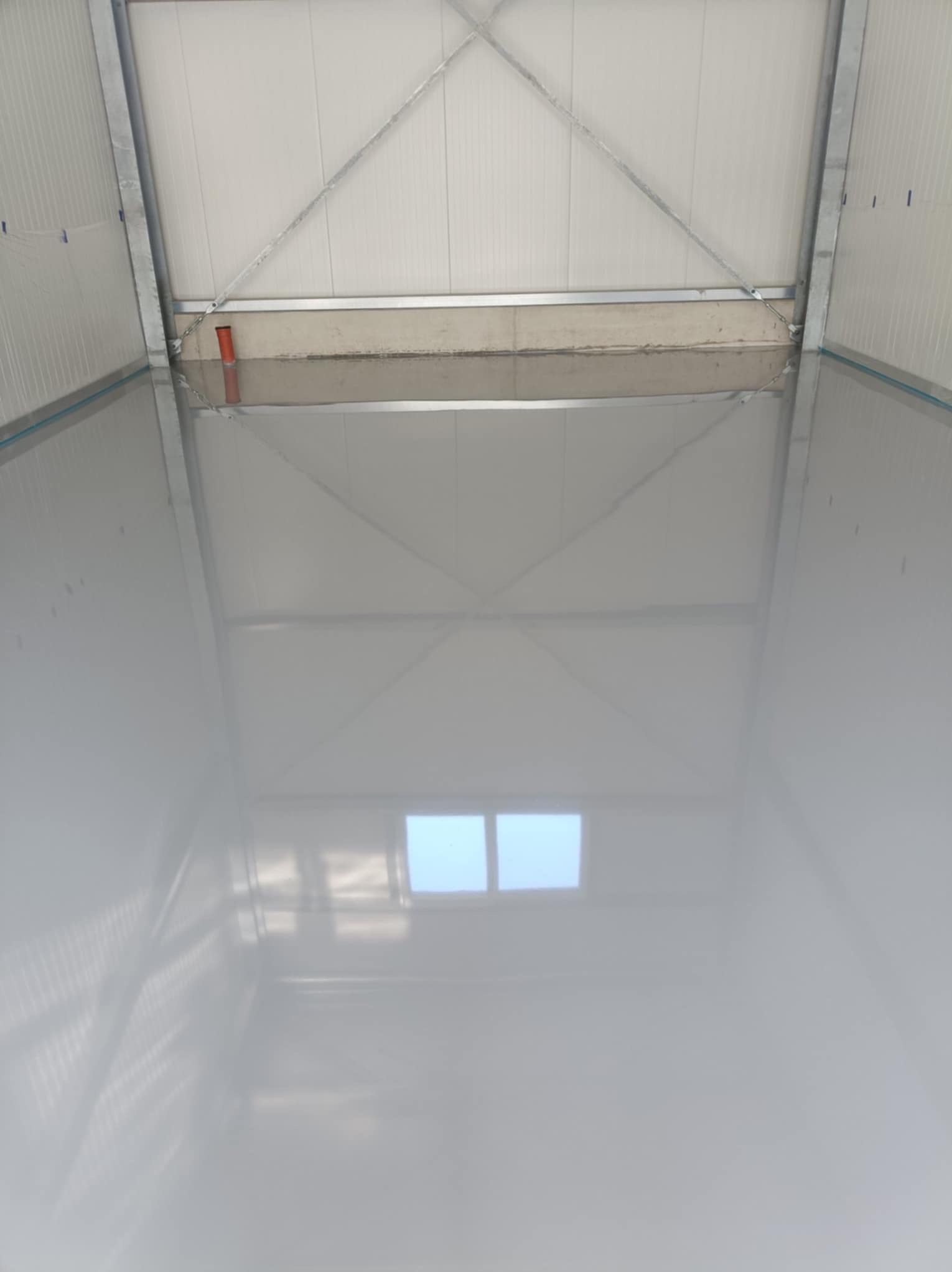 Błyszcząca, jasnoszara posadzka w hali magazynowej w Goleniowie, widziana z góry. Widoczne metalowe elementy konstrukcyjne dachu i ścian. W posadzce odbija się okno.