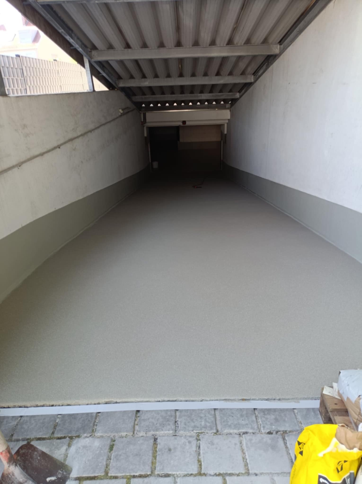 Jasnoszara posadzka żywiczna w garażu podziemnym, świeżo wylana, z widocznymi narzędziami i materiałami budowlanymi w tle. Gładka powierzchnia, równomierne oświetlenie.