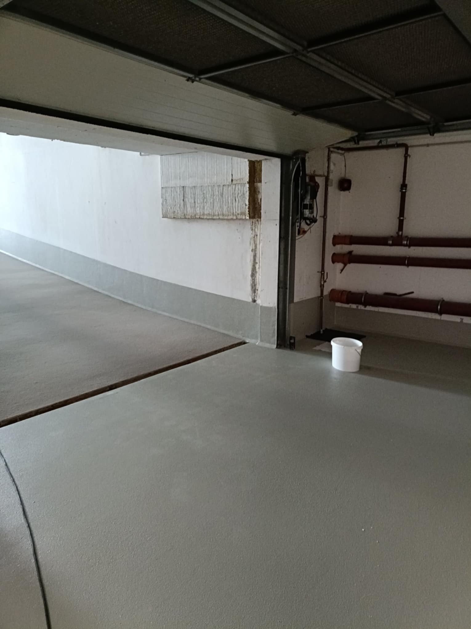 Jasnoszara, gładka posadzka żywiczna w garażu, z widocznym fragmentem progu i białym wiadrem. Na ścianie instalacja rurowa i fragment ocieplenia.