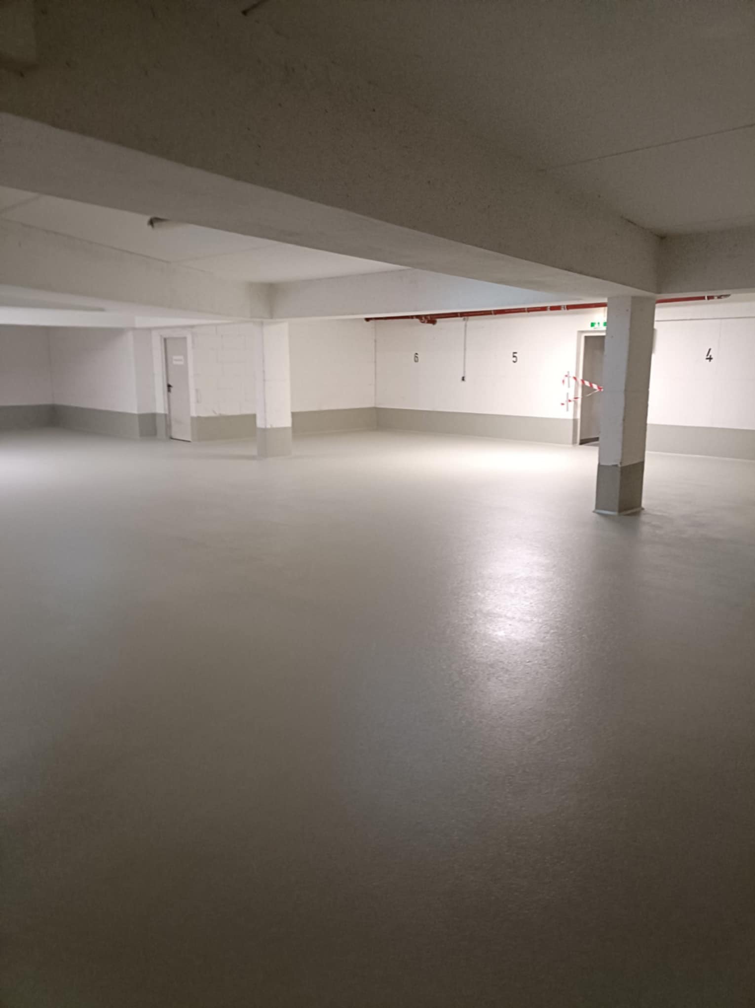 Jasnoszara, gładka posadzka w przestronnym, pustym pomieszczeniu z betonowymi filarami i numerami na ścianach. Widoczne drzwi ewakuacyjne.