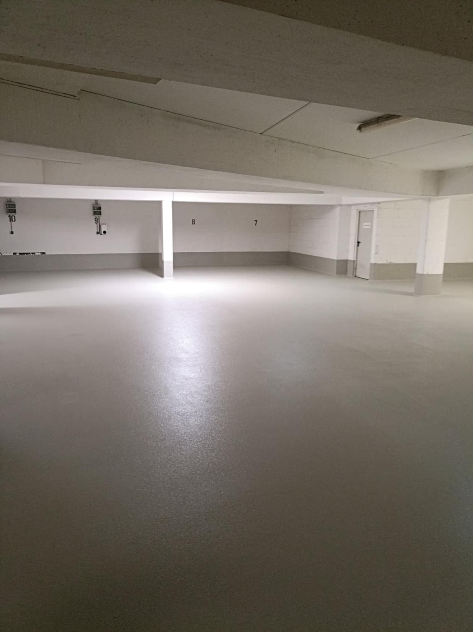Jasnoszara, gładka posadzka w garażu podziemnym z numerowanymi miejscami parkingowymi. Widoczne słupy i fragment drzwi.