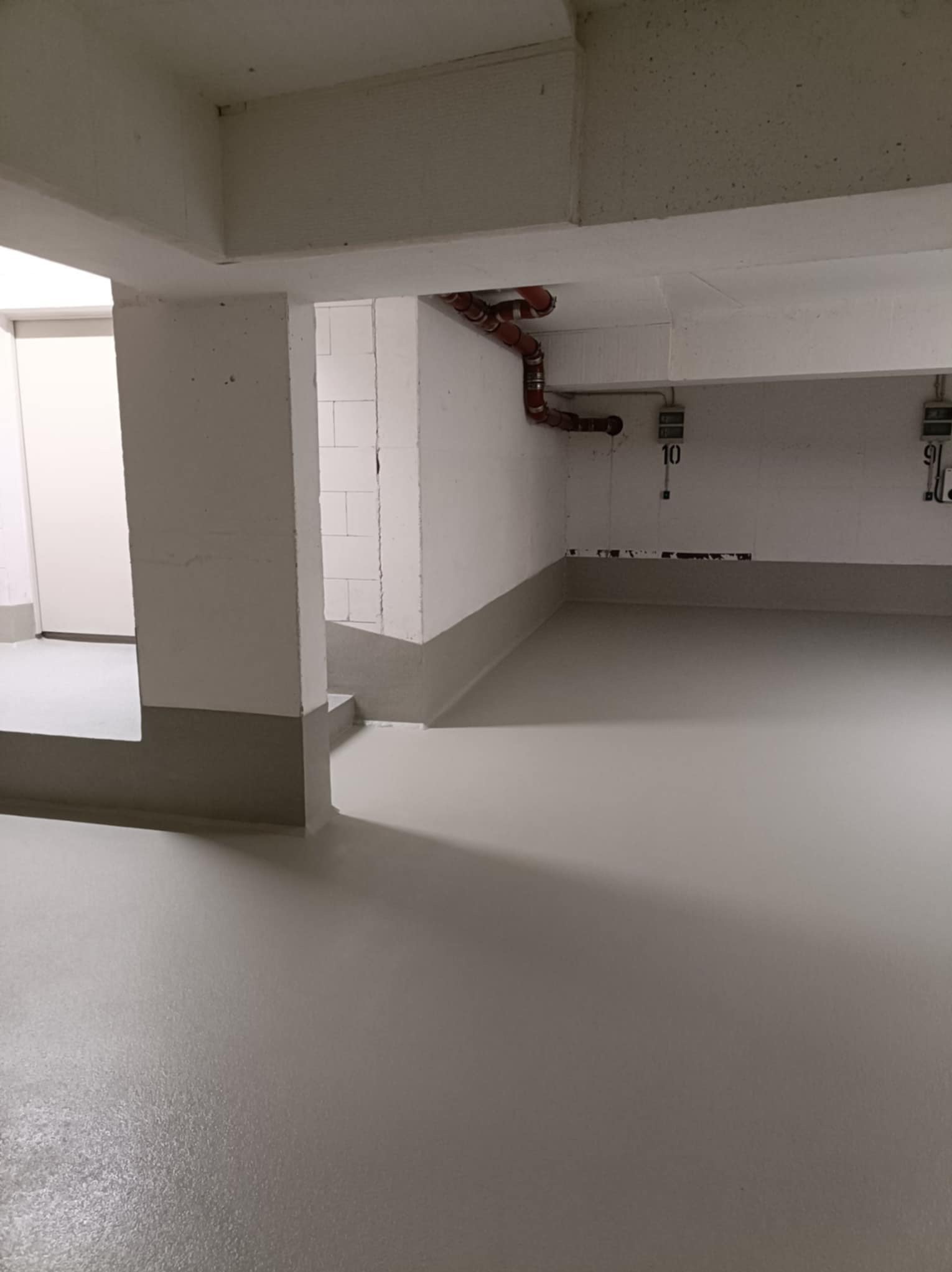 Jasnoszara, gładka posadzka w pomieszczeniu z białymi ścianami i widoczną instalacją. Minimalistyczne wnętrze z elementami technicznymi.