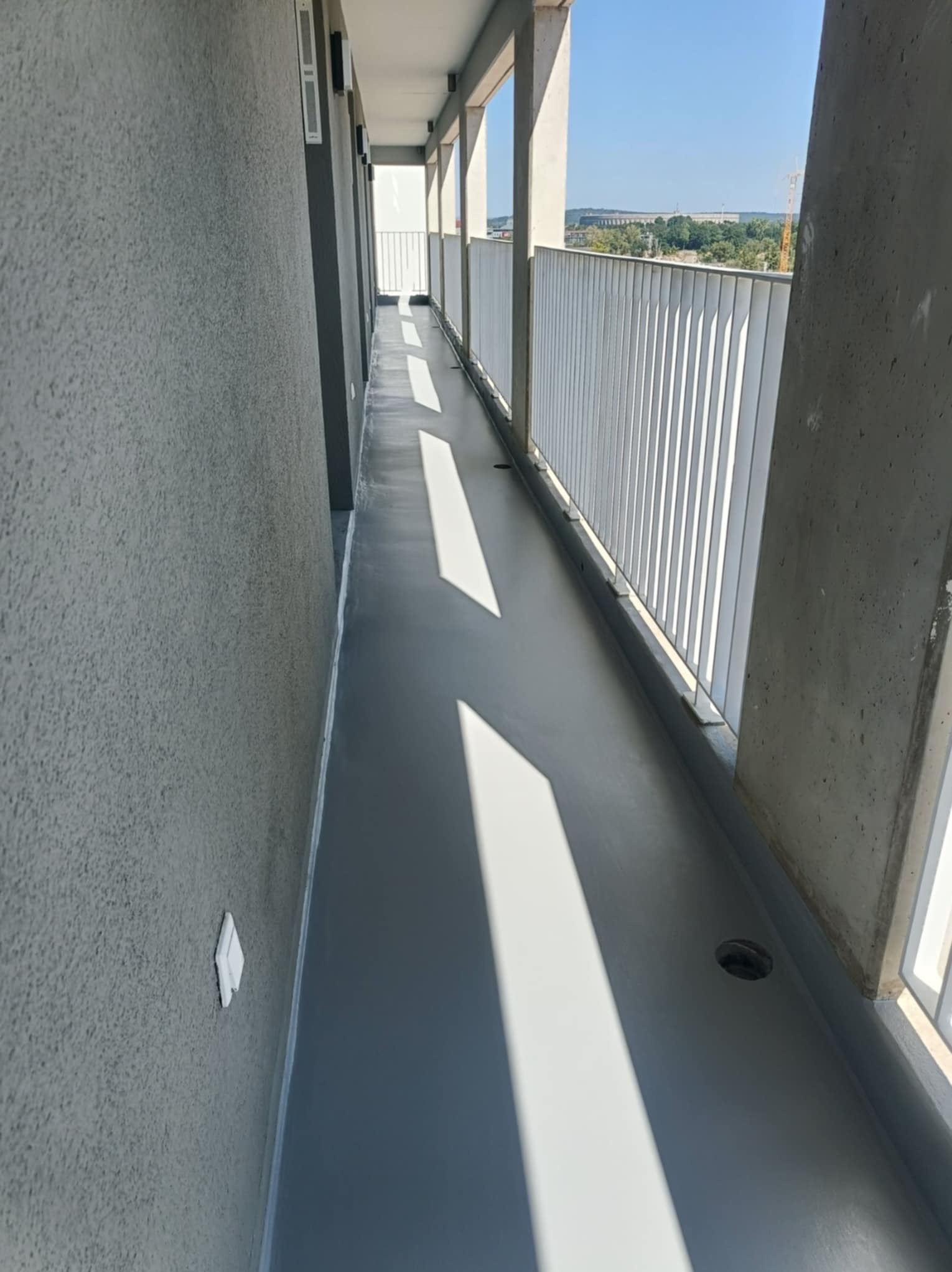 Długi balkon z szarą, gładką posadzką żywiczną, białą balustradą i widokiem na miasto w oddali. Mocne światło słoneczne tworzy geometryczne cienie na podłodze.