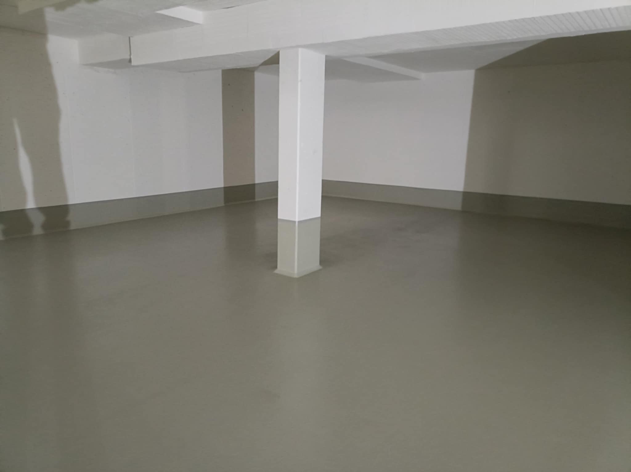 Gładka, szara posadzka w pustym pomieszczeniu z białymi ścianami i filarem. Minimalistyczne wnętrze, świeżo wylana podłoga, widoczne refleksy światła.