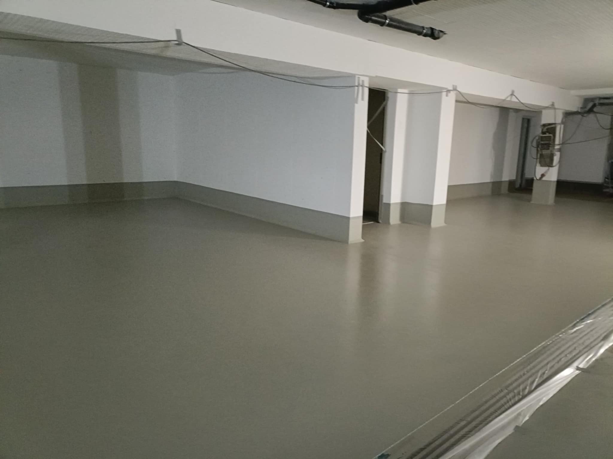 Jasnoszara, gładka posadzka w przestronnym, surowym wnętrzu z białymi ścianami i szarymi cokołami. Widoczne instalacje pod sufitem i fragment zabezpieczonej krawędzi podłogi.