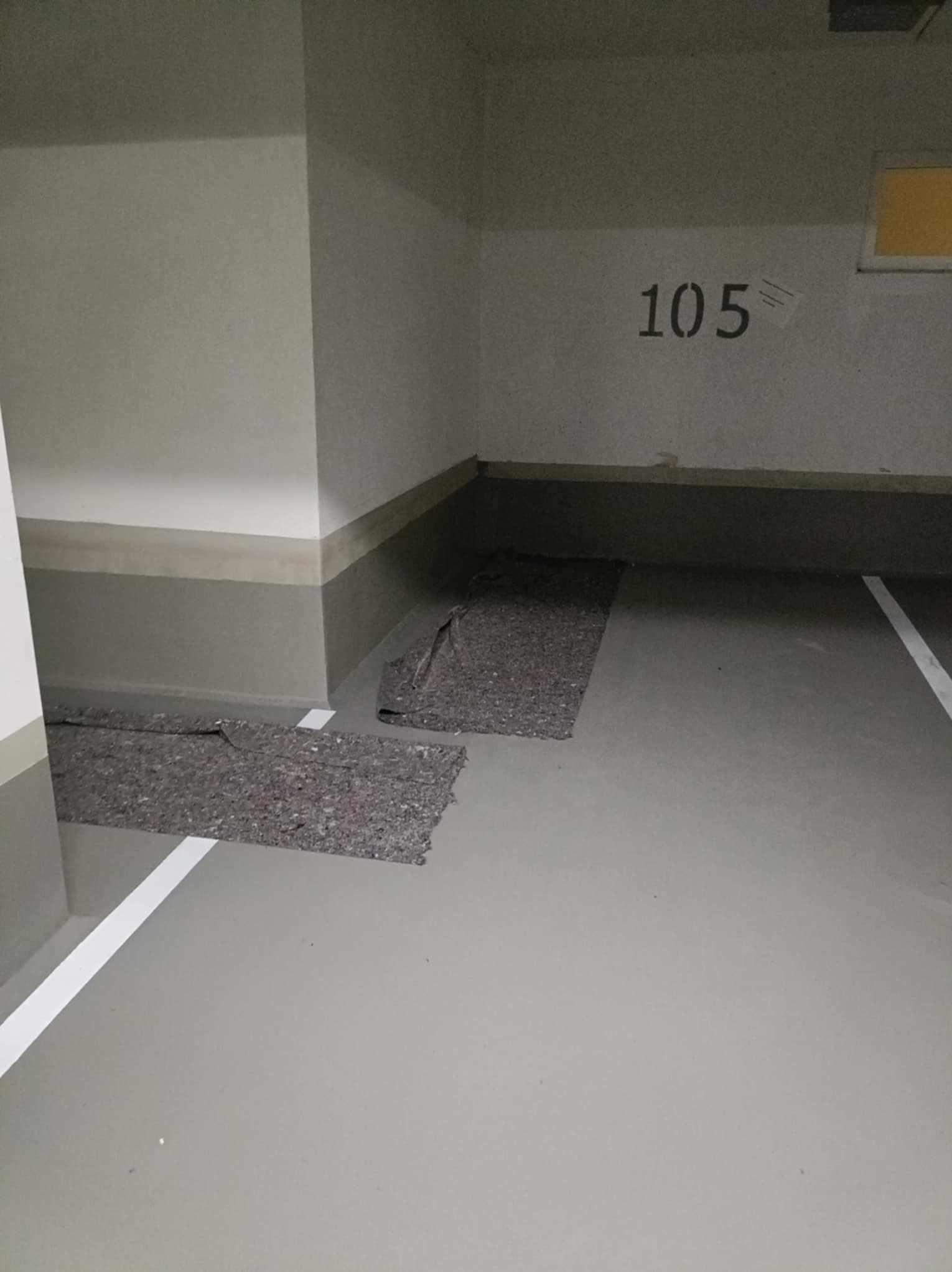 Szara posadzka w garażu z numerem 105 na ścianie, częściowo zakryta szarym materiałem. Widoczne białe linie wyznaczające miejsca parkingowe.