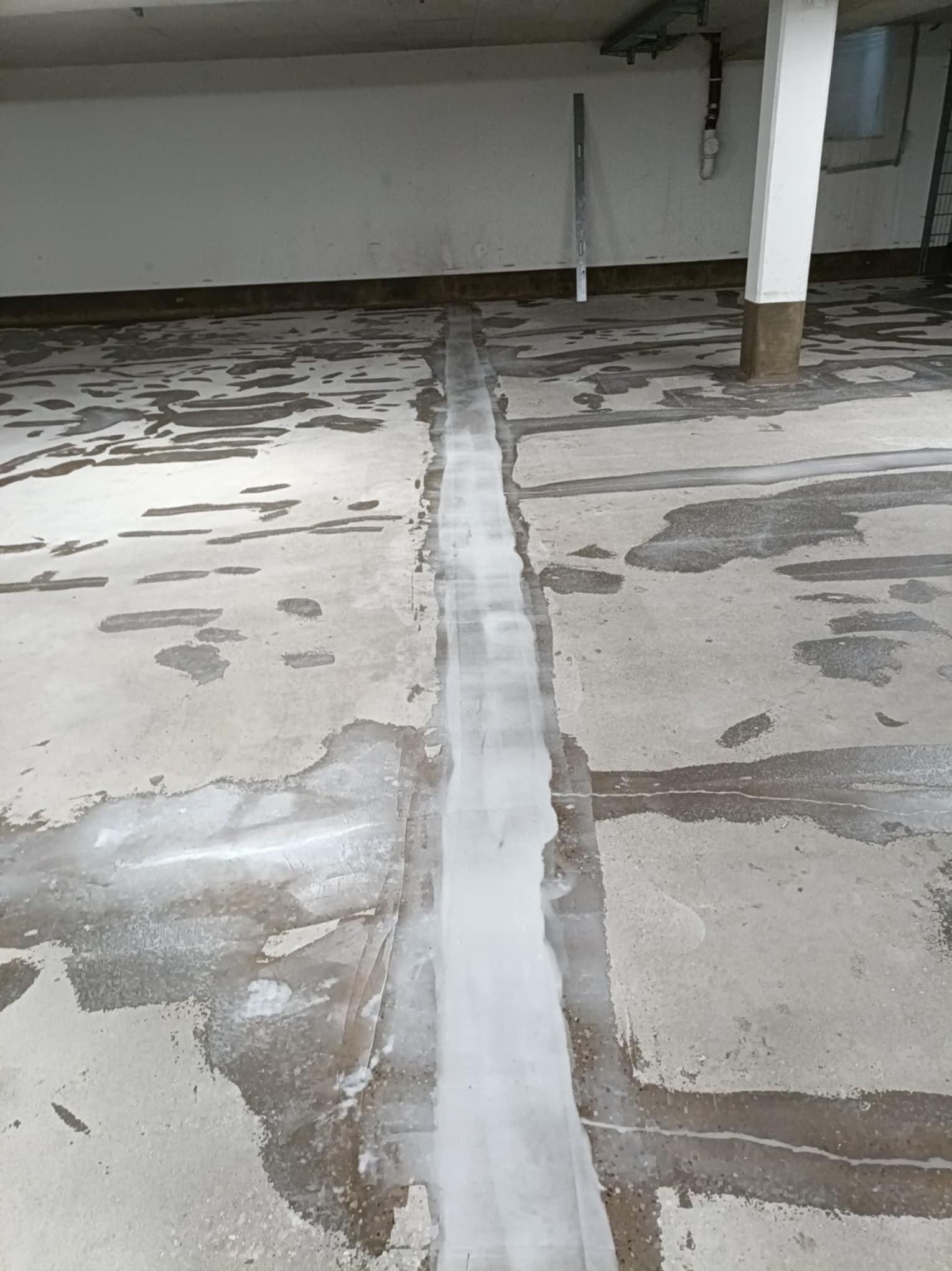 Przygotowanie podłoża pod posadzkę żywiczną: gruntowanie szczelin dylatacyjnych w szarej, betonowej podłodze garażu. Widoczny poziomica i filar.