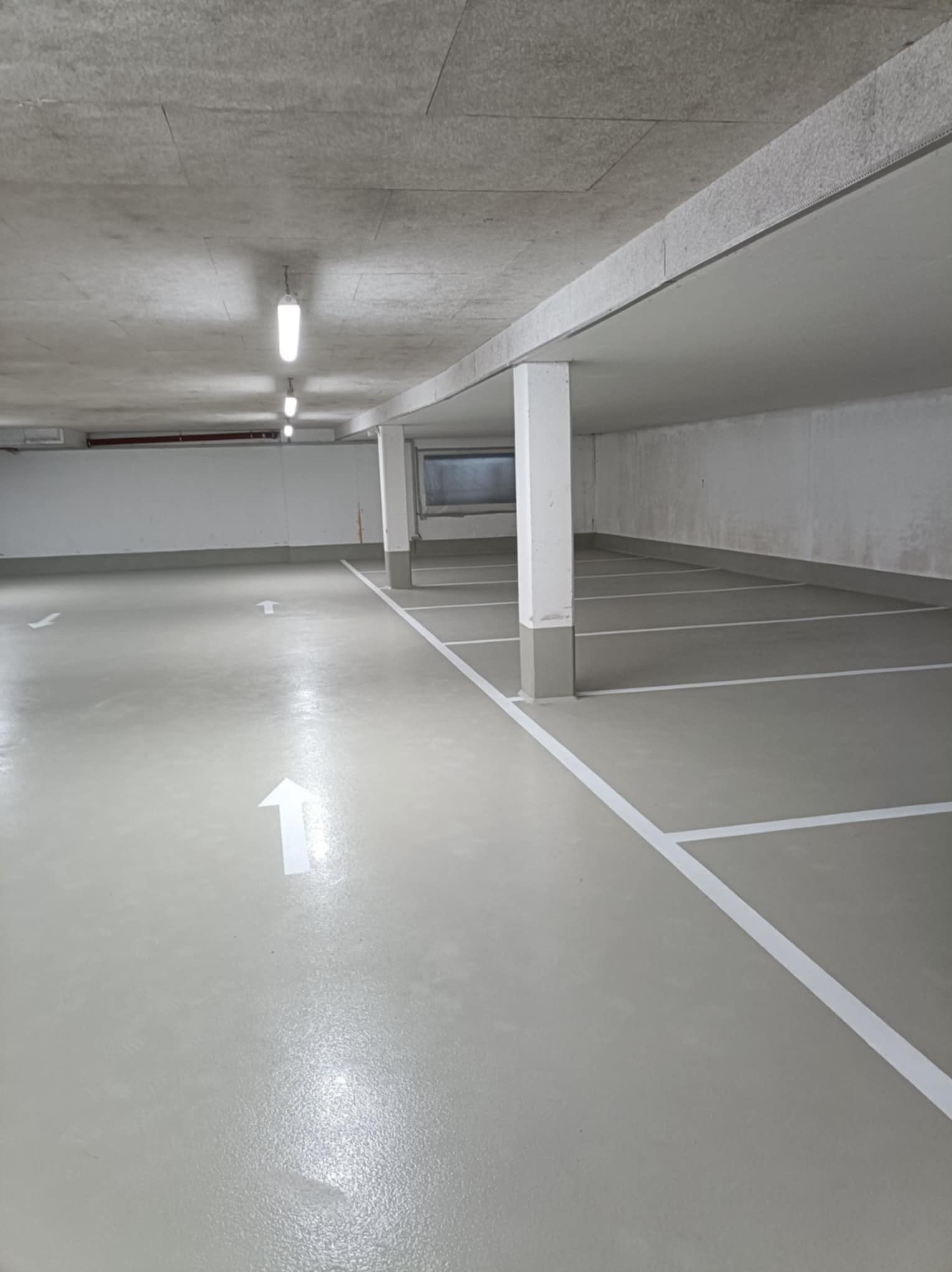 Jasna posadzka w garażu podziemnym z białymi liniami wyznaczającymi miejsca parkingowe i strzałkami kierunkowymi. Widoczne kolumny i oświetlenie sufitowe.