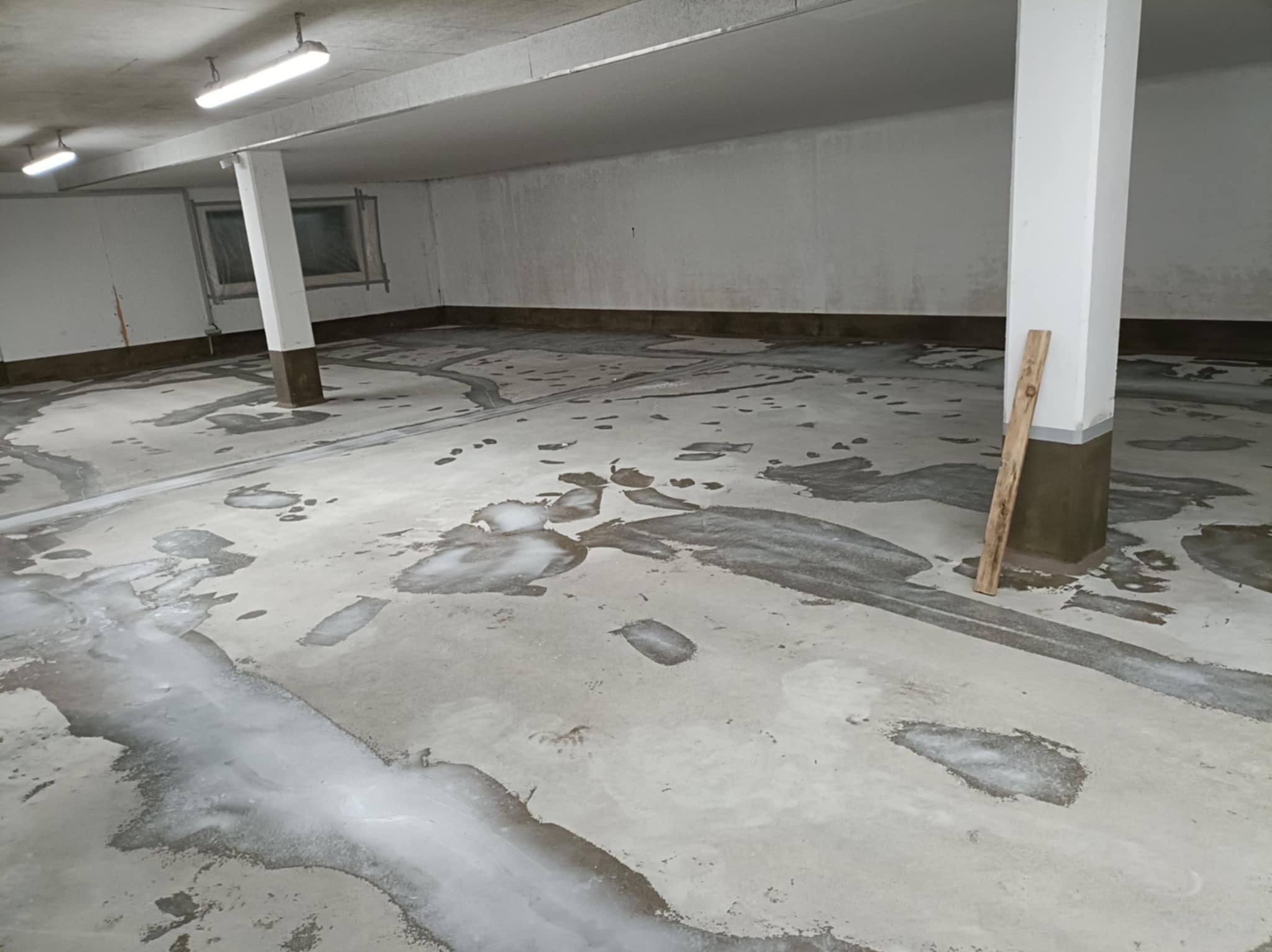 Przygotowanie podłoża pod posadzkę żywiczną: gruntowanie w garażu z kolumnami. Widoczne ślady mokrego gruntu i drewniana listwa oparta o kolumnę.