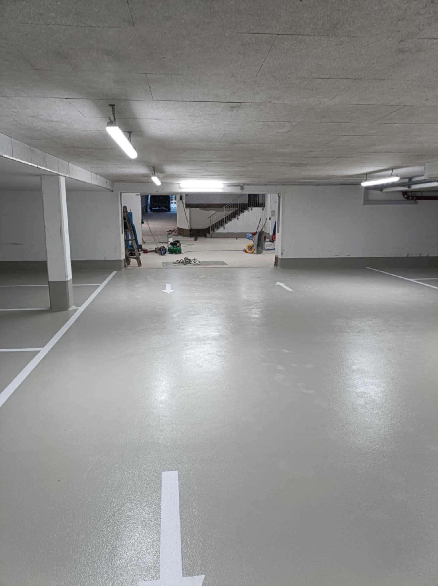 Jasnoszara posadzka w garażu podziemnym z wymalowanymi białymi liniami i strzałkami kierunkowymi. W tle widoczne narzędzia i schody.