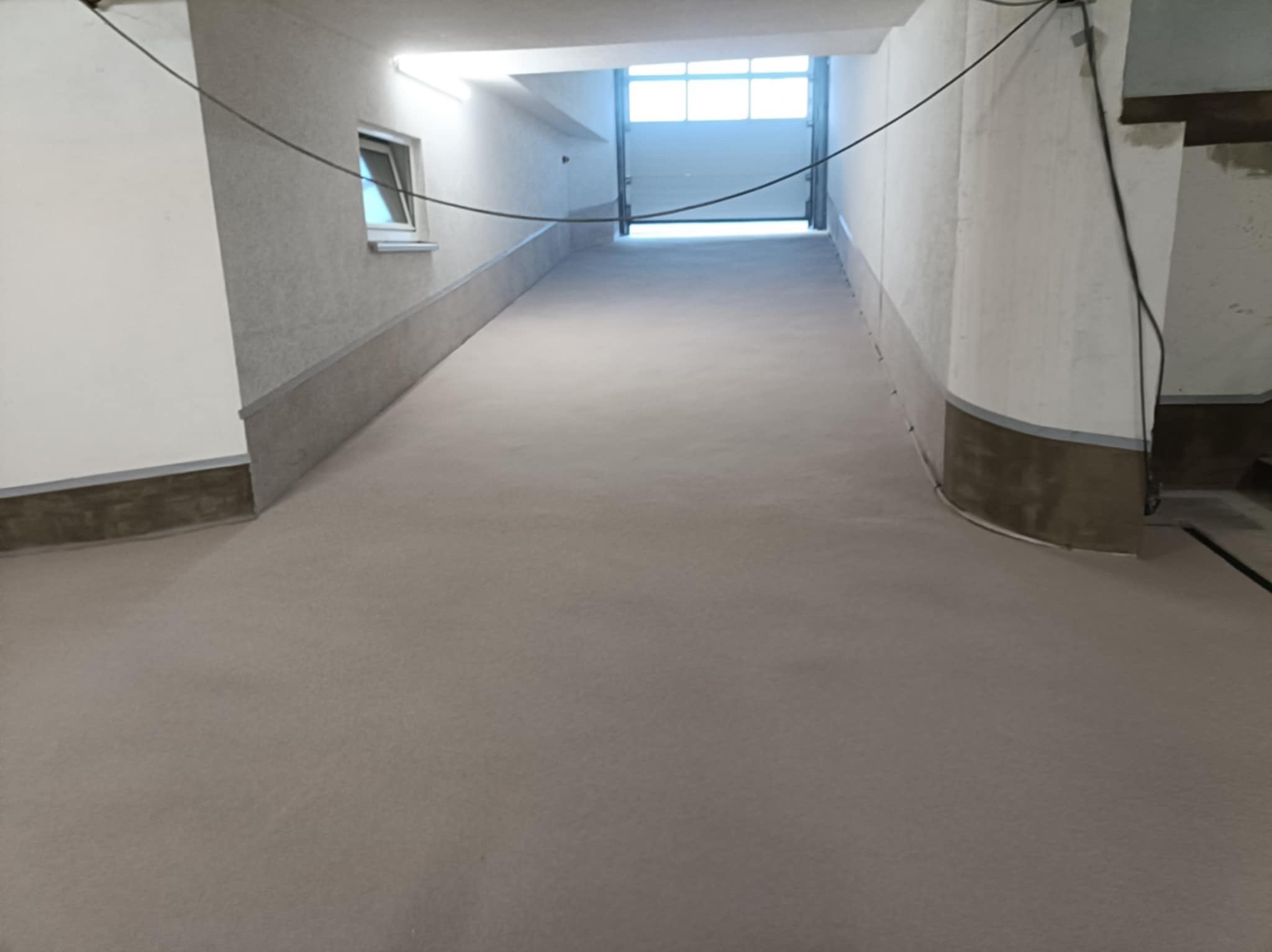 Gładka, szara posadzka w korytarzu z oknem i bramą garażową w tle. Widoczne ściany z szarym cokołem. Minimalistyczny styl, surowe wykończenie.