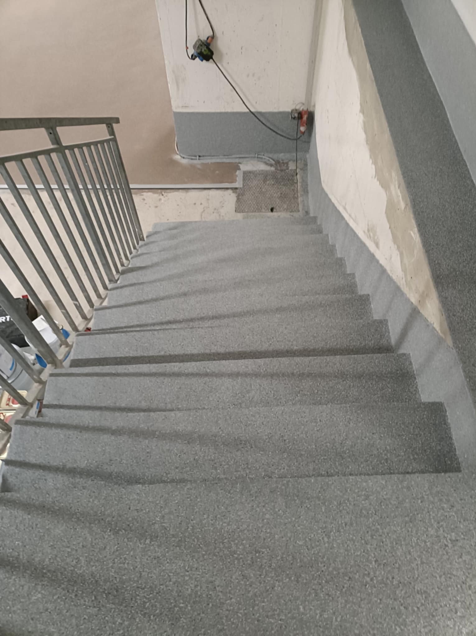Schody w szarym kolorze z żywicy epoksydowej, wykończone listwami przypodłogowymi. Metalowa barierka po lewej stronie. Widok z góry na dół.