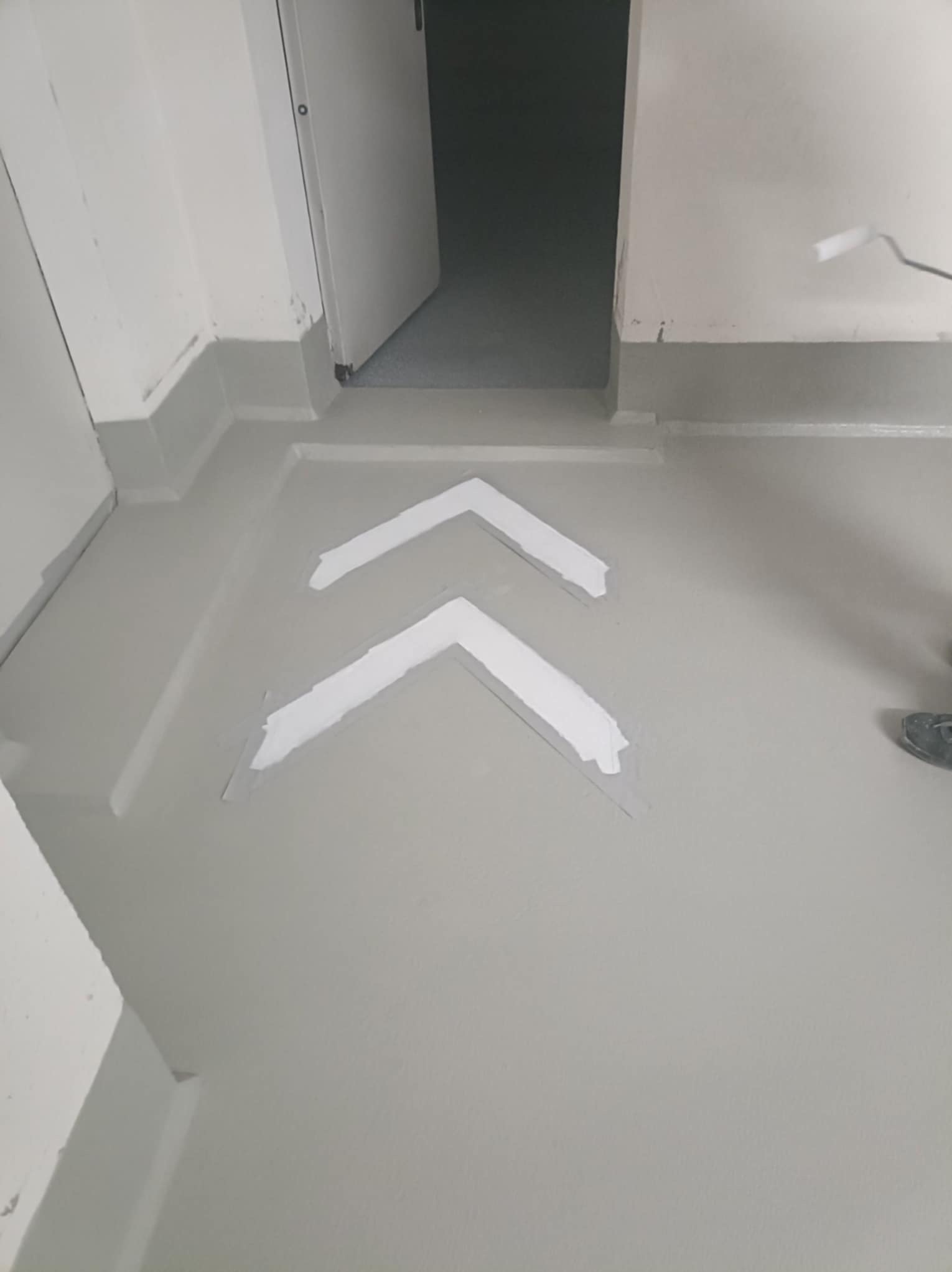 Szara posadzka w korytarzu z białymi strzałkami kierunkowymi, malowana wałkiem przy ścianie. Widoczne drzwi i fragment buta malarza.