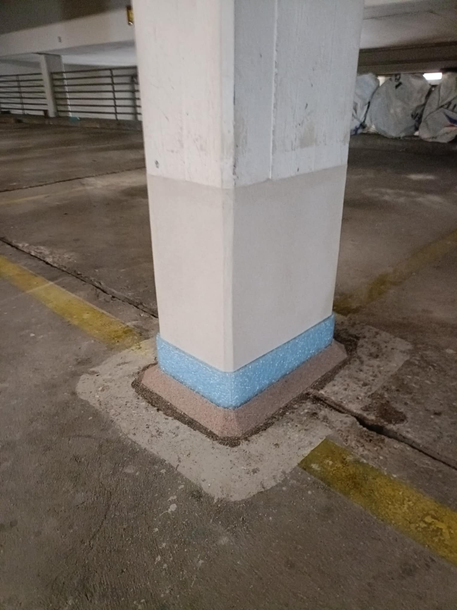 Ochrona słupa w garażu wielopoziomowym: obudowa z brązowych i niebieskich elementów, zabezpieczająca przed uszkodzeniami. Żółta linia na posadzce.