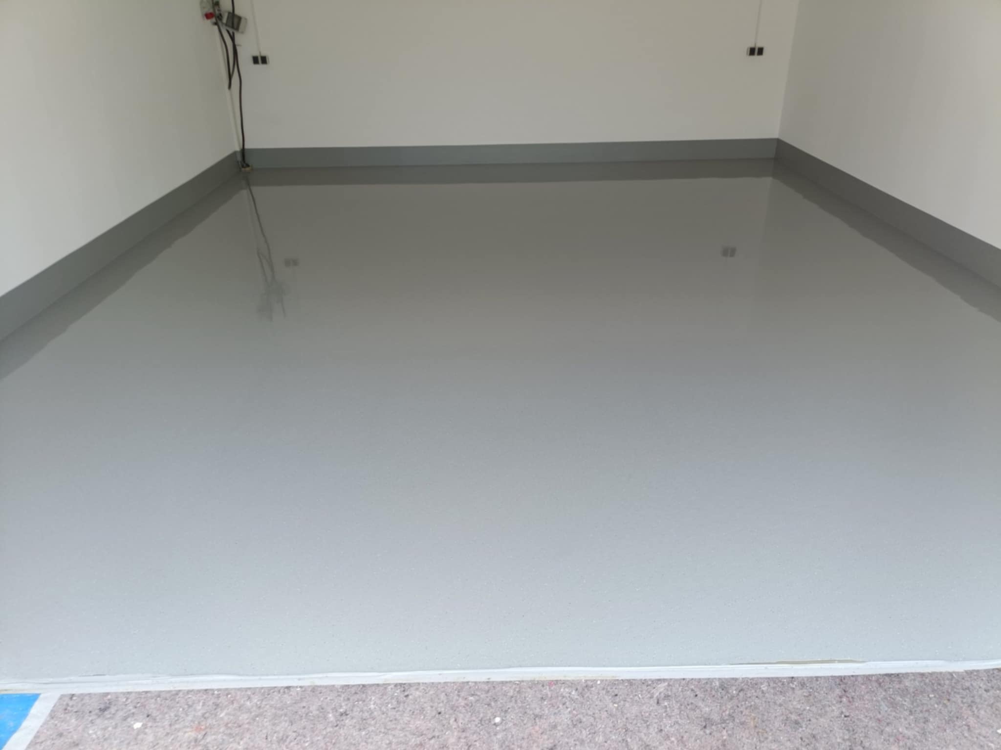 Jasnoszara, gładka posadzka żywiczna w pomieszczeniu z białymi ścianami i szarymi listwami przypodłogowymi. Widoczne kable elektryczne przy suficie.